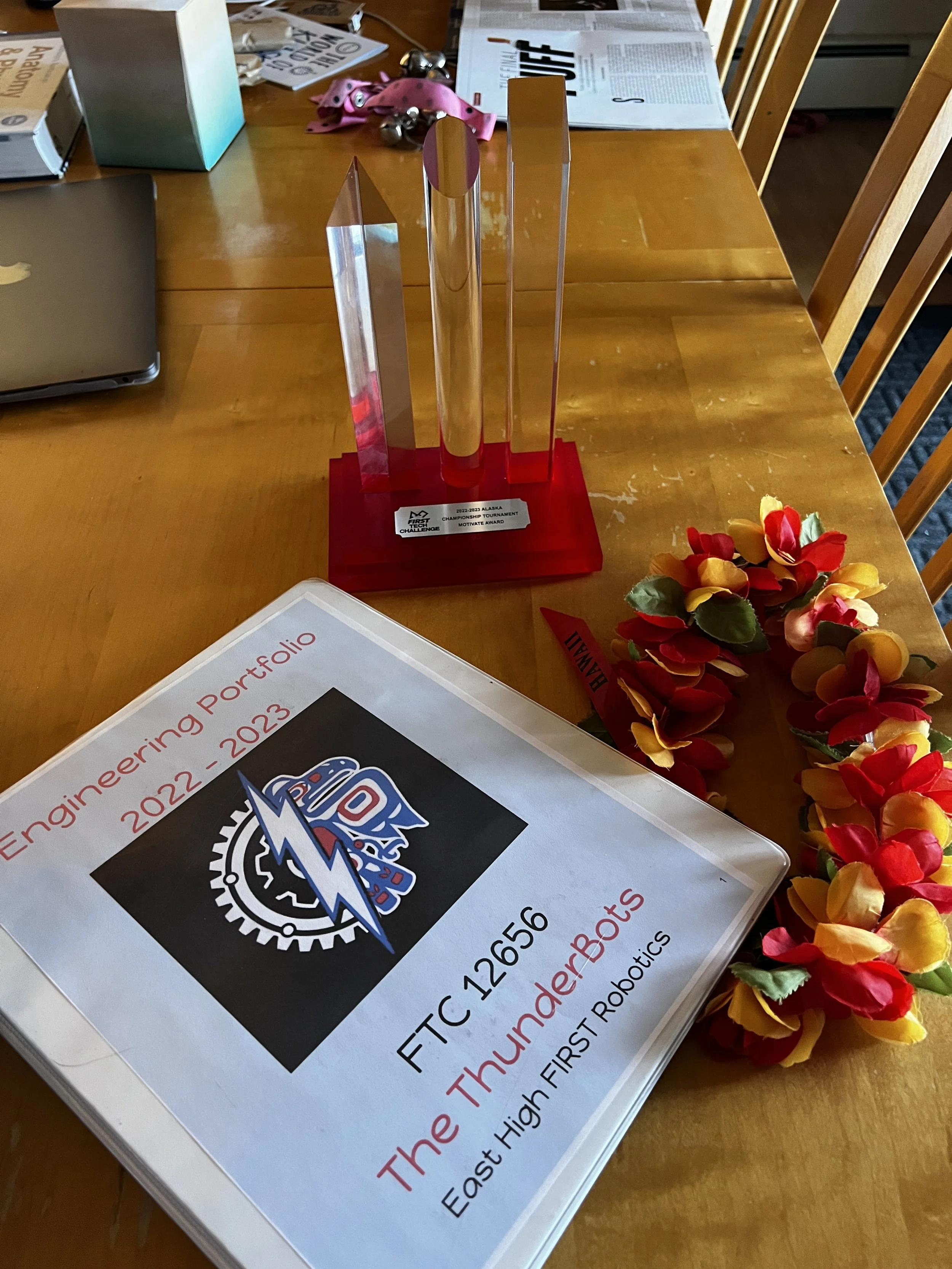 A wooden table with a first-place trophy, a diploma for engineering team, a floral headband, and other papers and objects.
