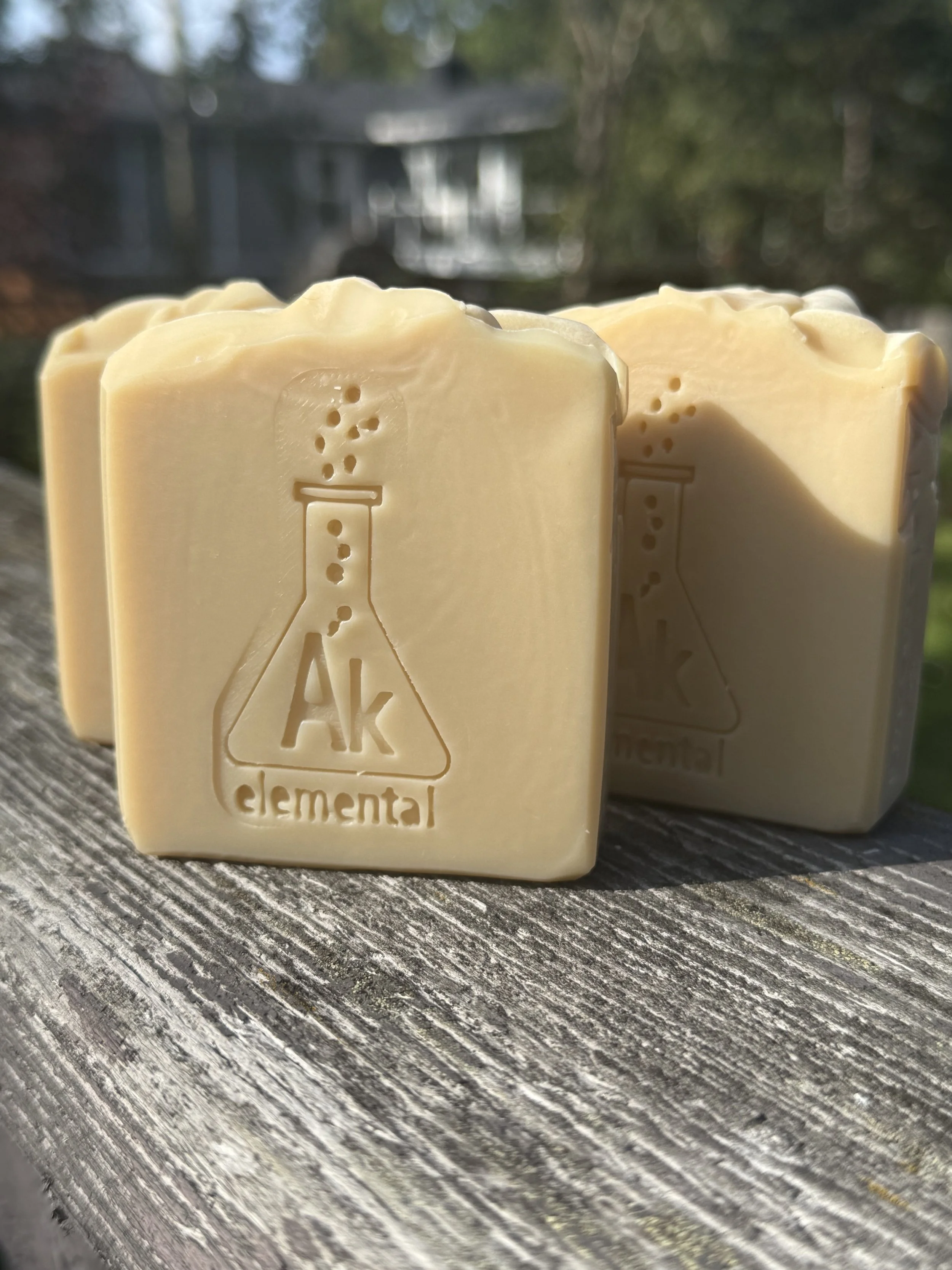 Three creamy, uncolored Sugar Cookie handcrafted soap bars with an embossed AK Elemental flask logo, displayed on rustic wood outdoors.