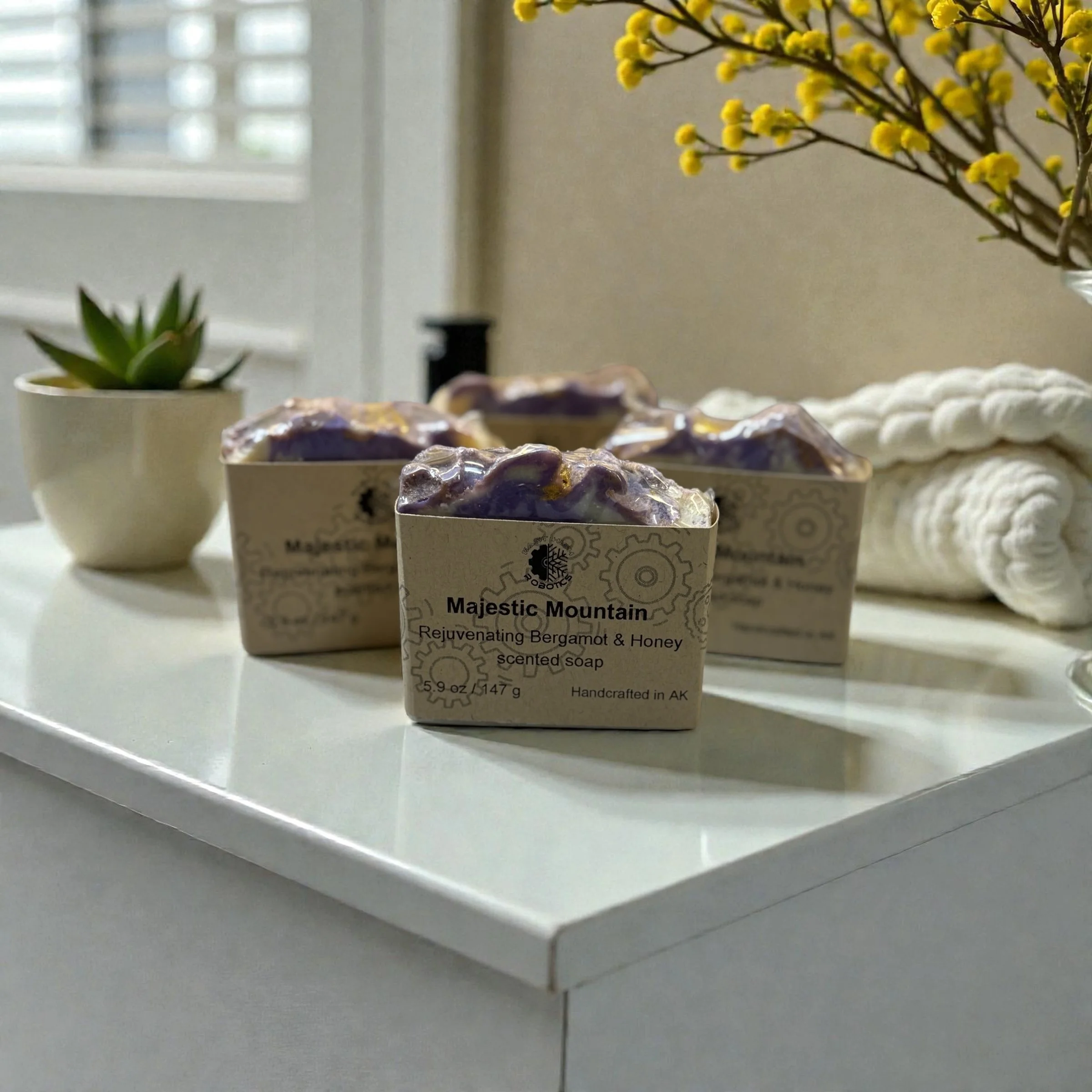Three soaps sitting on a counter in a bathroom. The soaps have purple tops and are wrapped in brown labels with the text Majestic Mountain.