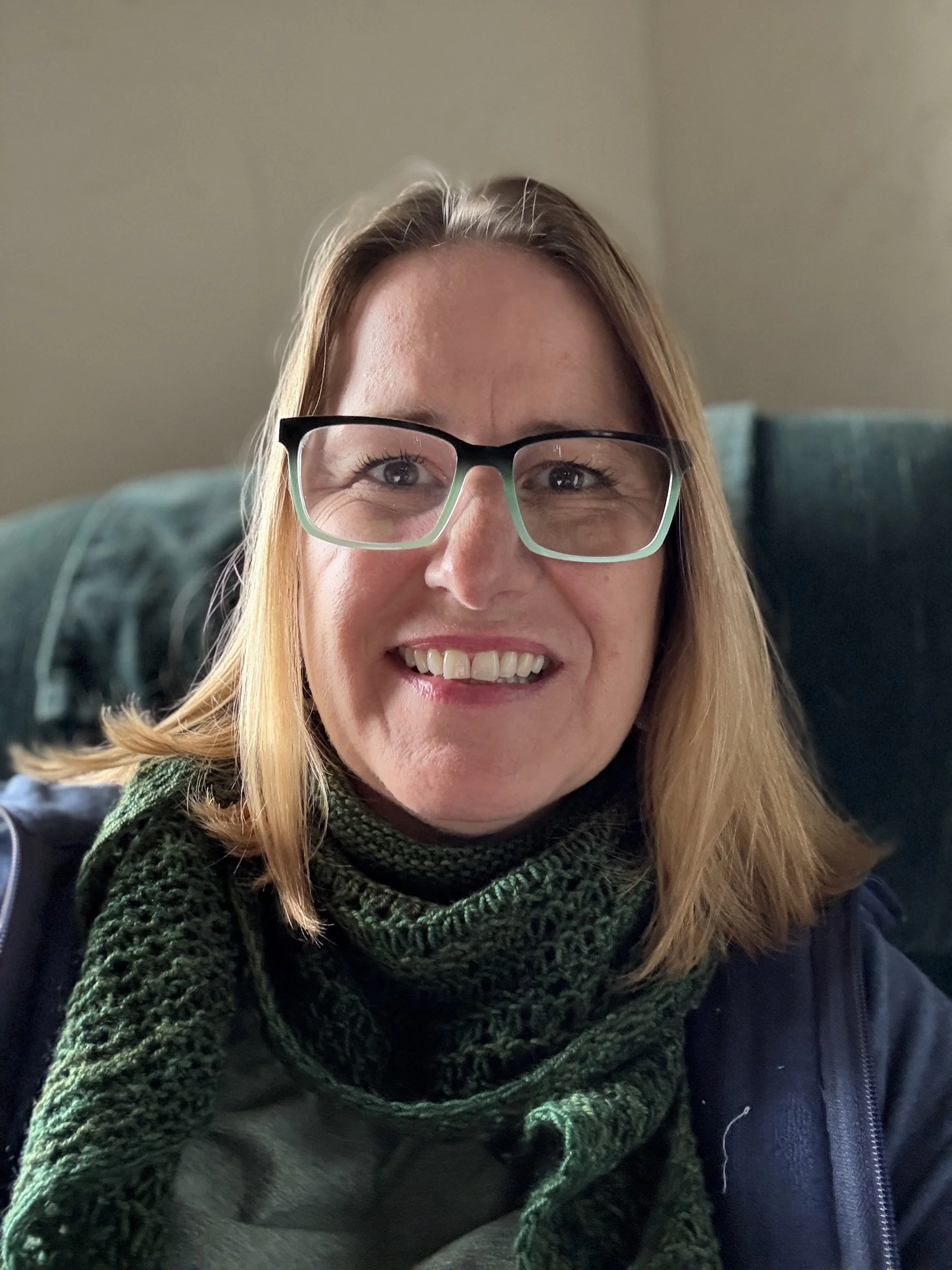 A woman with glasses and blonde hair smiling at the camera, sitting on a green couch.