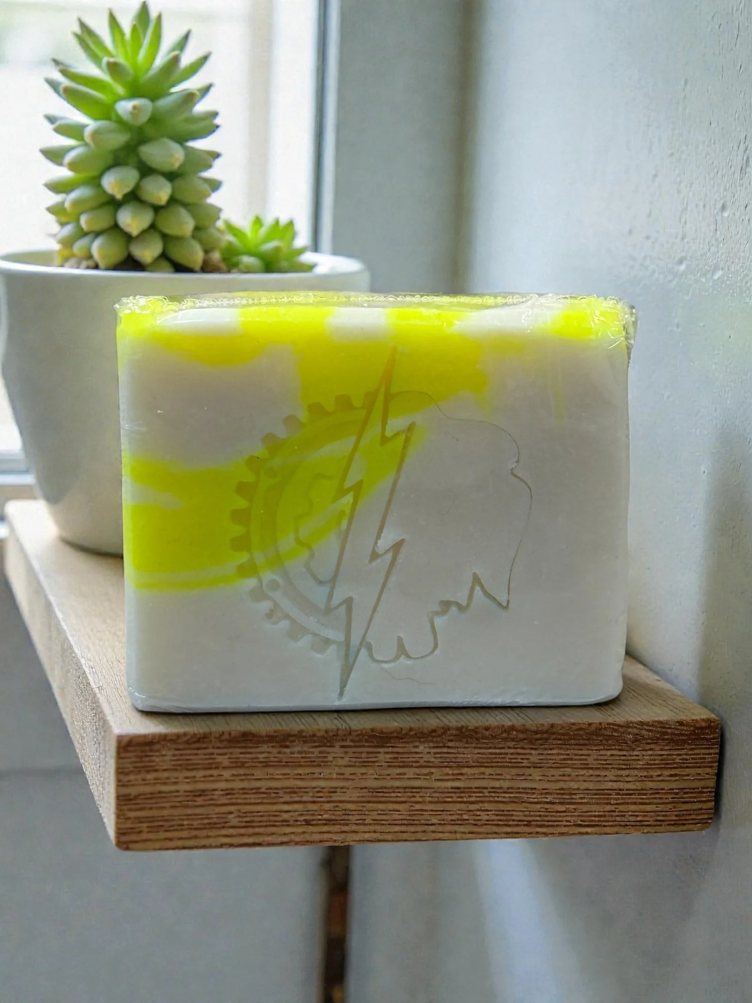 Single bar of yellow & white soap sitting on a wooden shelf with a succulent plant in the background. Soap has a stamp impression in the shape of a gear.