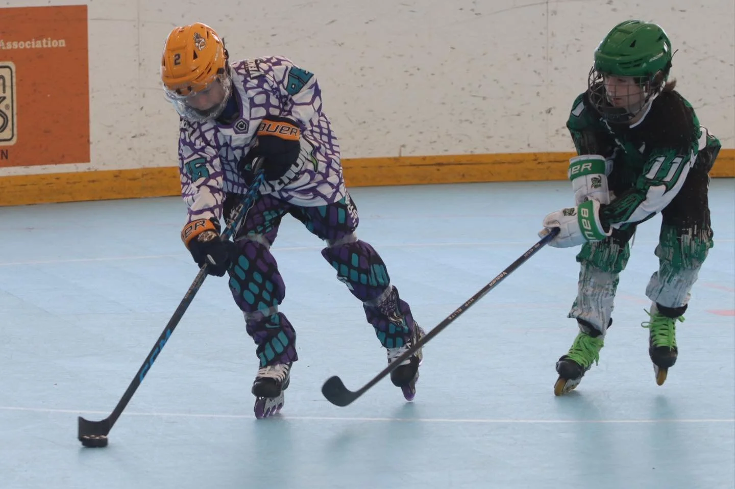 Two children playing roller hockey on an indoor rink, wearing colorful jerseys, helmets, and protective gear, competing for the puck.