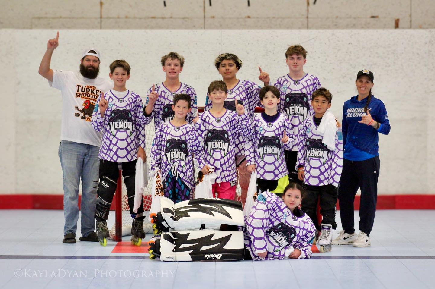 A youth hockey team in purple and black jerseys with the word "VENOM" on them, posing with two coaches or trainers in a rink. The children are holding hockey sticks and wearing protective gear, with a goalie kneeling in front. The team is smiling and pointing upwards, with some players making trumpet gestures. The background shows an indoor rink wall.
