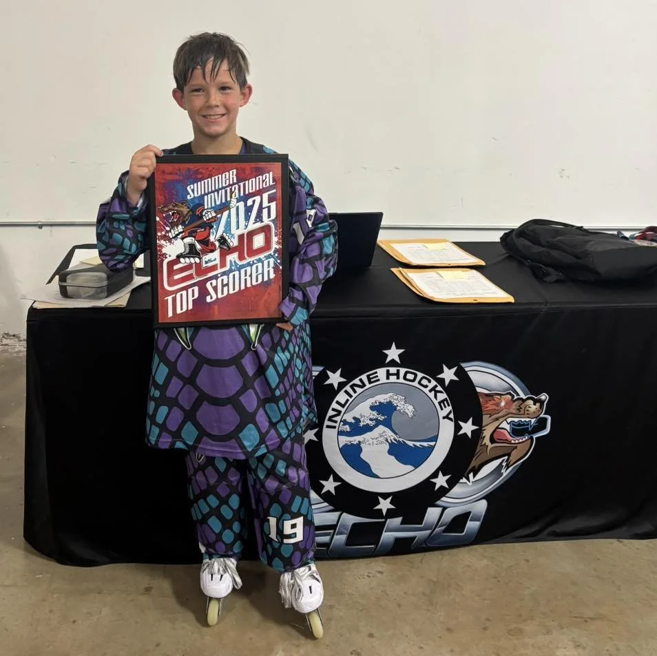 A young boy in colorful hockey gear, wearing roller skates, holding a sign that reads 'Summer Invitational 2025, 10, Top Scorer,' standing in front of a table with hockey-related logos and documents.
