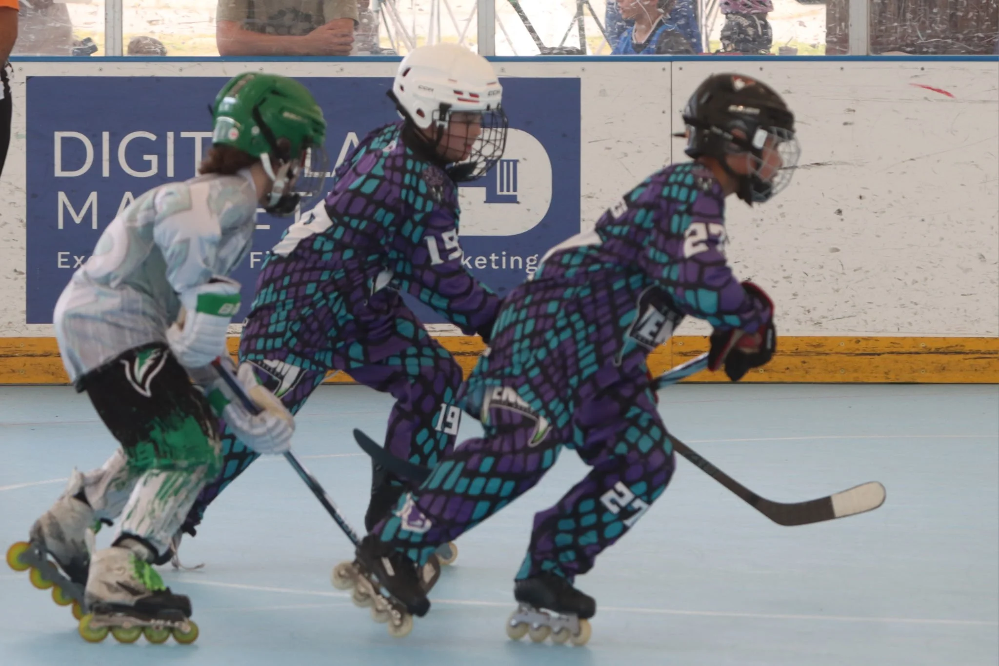 Three young children roller skating on an indoor rink, wearing helmets and colorful outfits with purple, teal, and white patterns, skating in a line.