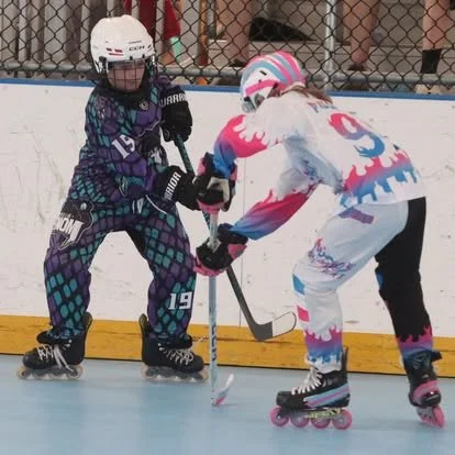 Two young girls playing roller hockey on an outdoor rink, wearing helmets and protective gear, battling for control of the puck.