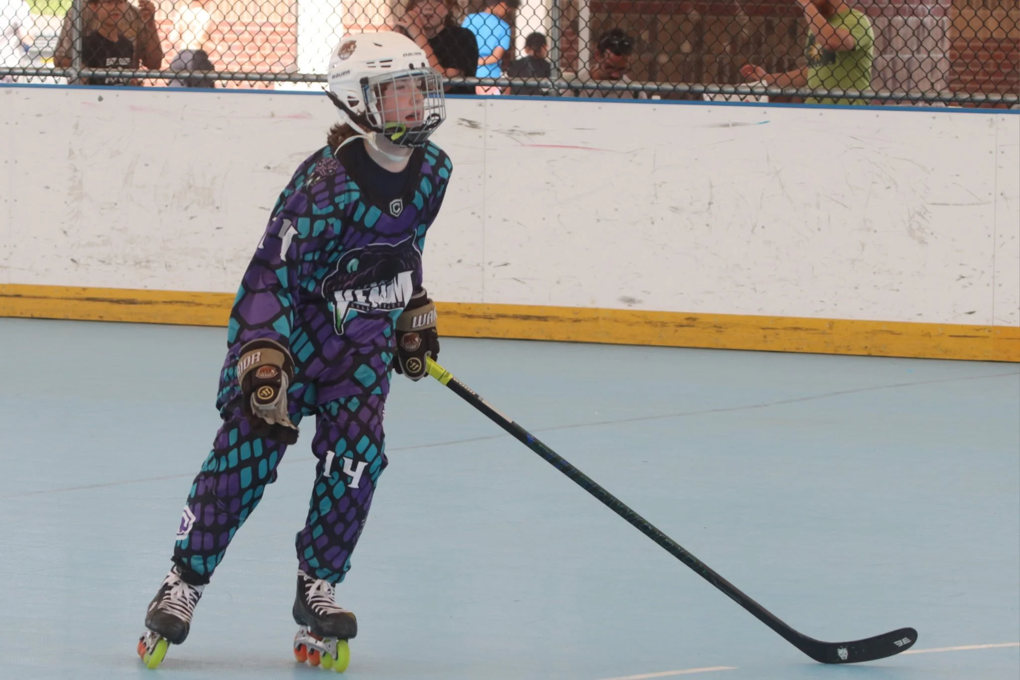 A young hockey player in colorful uniform skating on an inline hockey rink with a white helmet and holding a hockey stick.