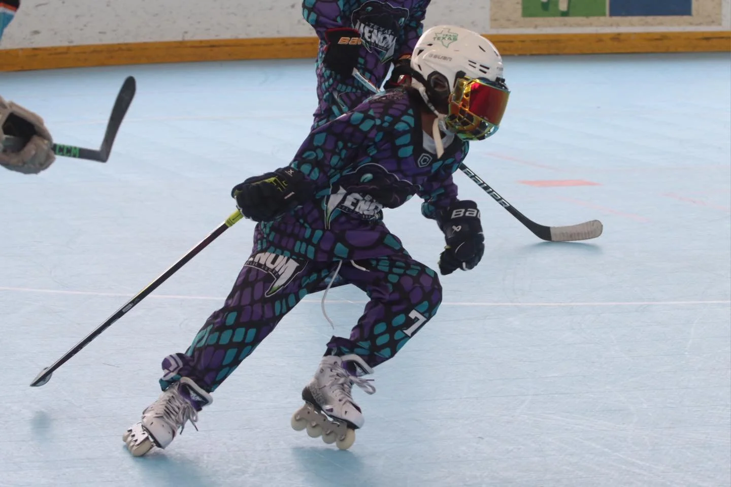 Young child in colorful hockey gear skating on an indoor rink, wearing a helmet with a red reflective visor, holding a hockey stick, and chasing a puck.