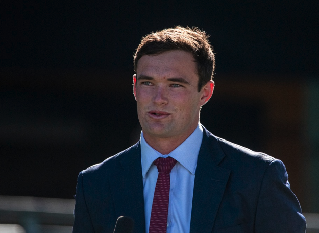 A young man in a navy blue suit and red tie standing outdoors, speaking into a microphone.