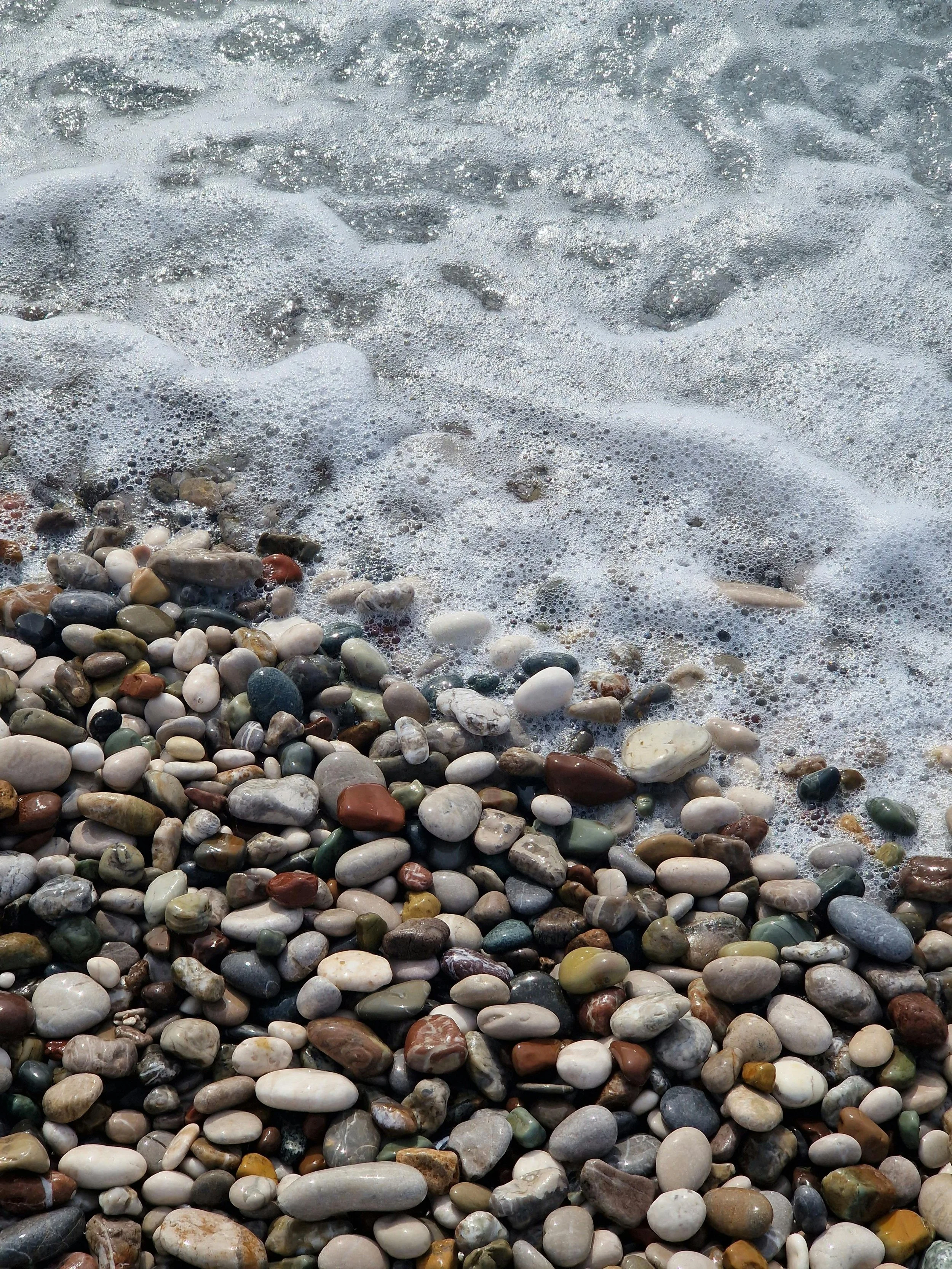 Close-up of colorful pebbles on a beach, with waves and foam approaching.