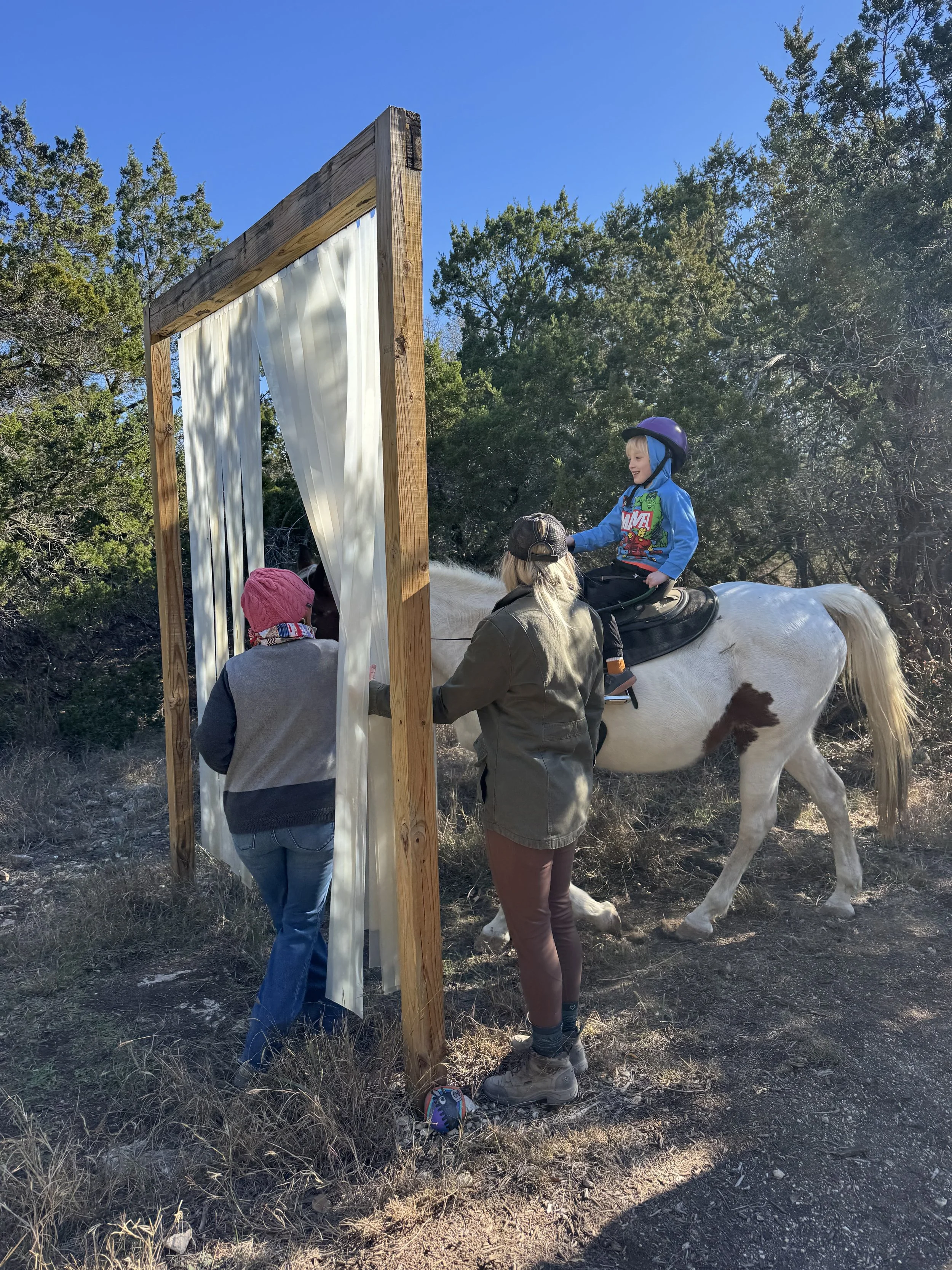 A young boy riding a white horse with brown spots approaches a makeshift outdoor curtain or barrier with two women nearby, in a wooded area with clear blue sky.