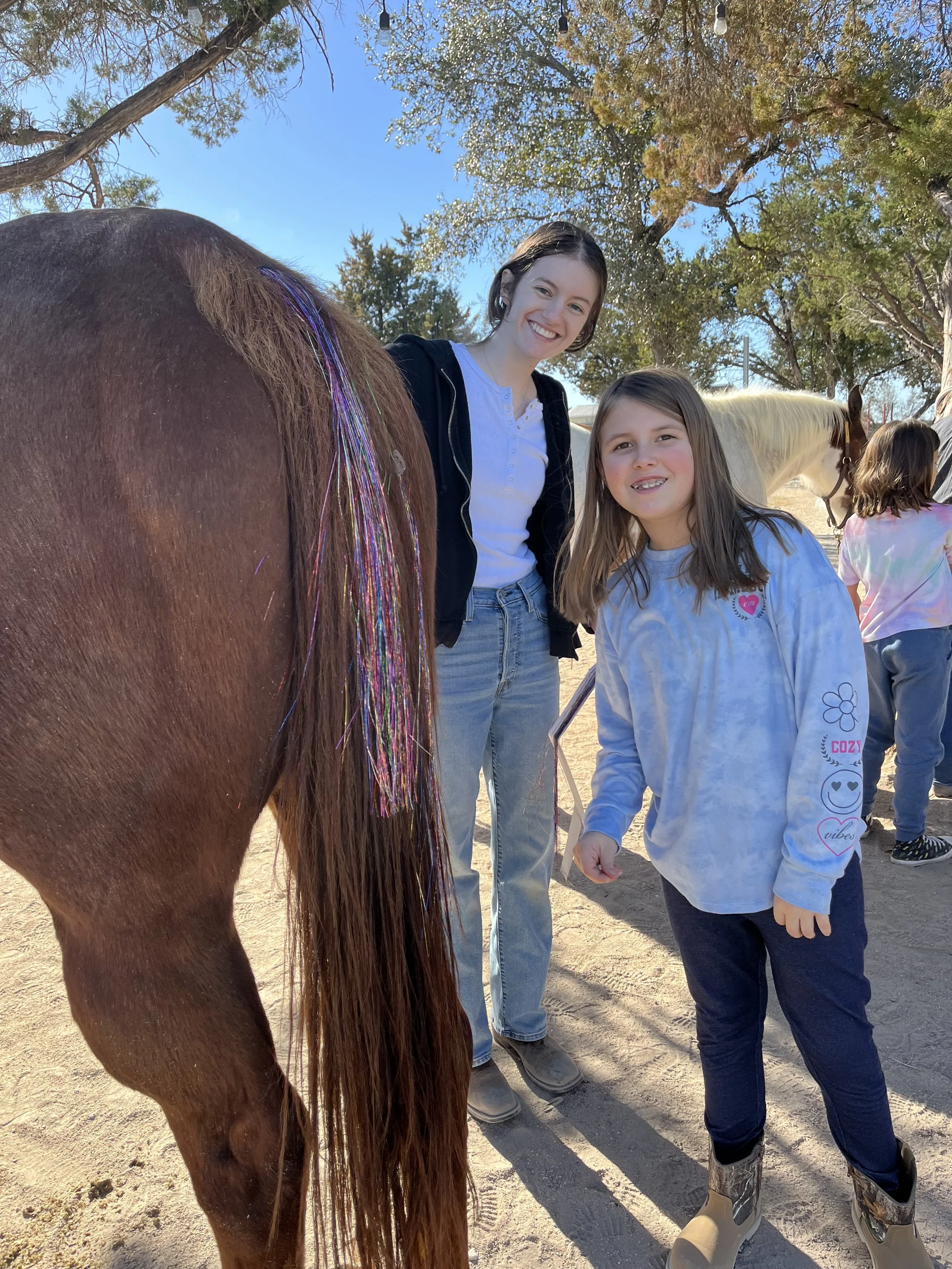 A woman and a girl standing outdoors near a brown horse with colorful hair extensions, smiling at the camera, with other children and horses in the background on a sunny day.