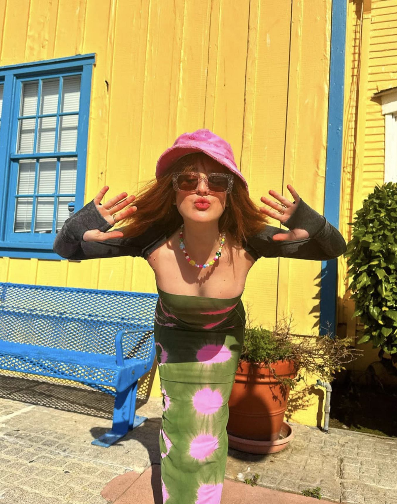 A woman dressed in colorful clothing, wearing sunglasses, a pink hat, and a beaded necklace, standing in front of a yellow building with blue window frames and a blue bench, making a playful gesture with her hands near her face.