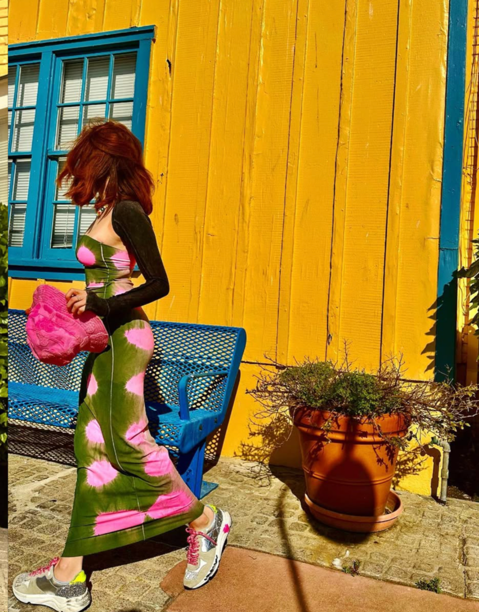 Woman in a long, colorful tie-dye dress holding a pink textured handbag, standing next to a blue metal bench and a large terracotta pot with a plant, against a bright yellow wooden wall with a blue window.