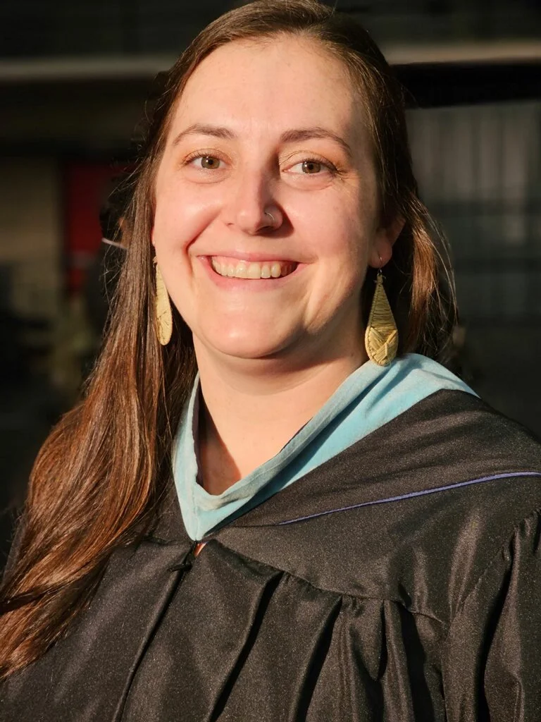 A woman with long brown hair smiling, wearing a graduation gown and earrings, with a blurred indoor background.