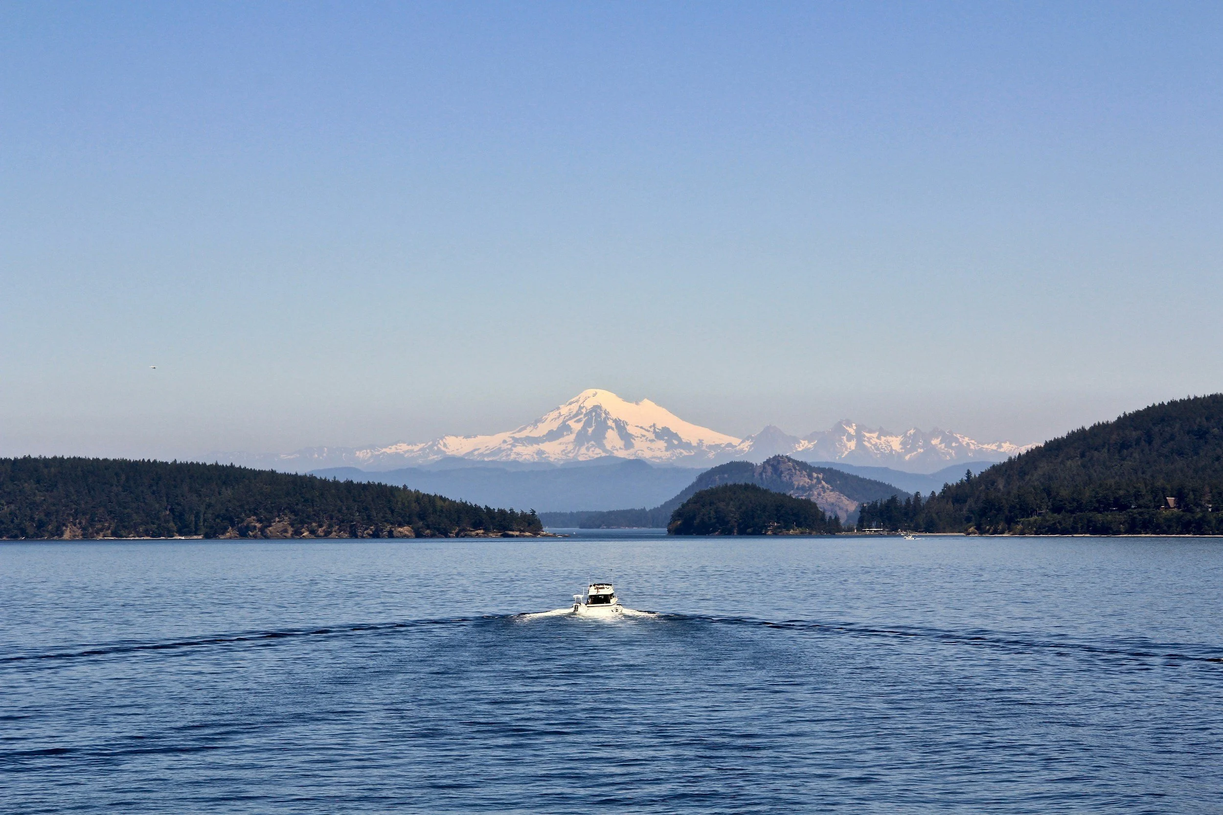 A boat on a large body of water with forested islands and mountains, including a snow-capped mountain in the background.