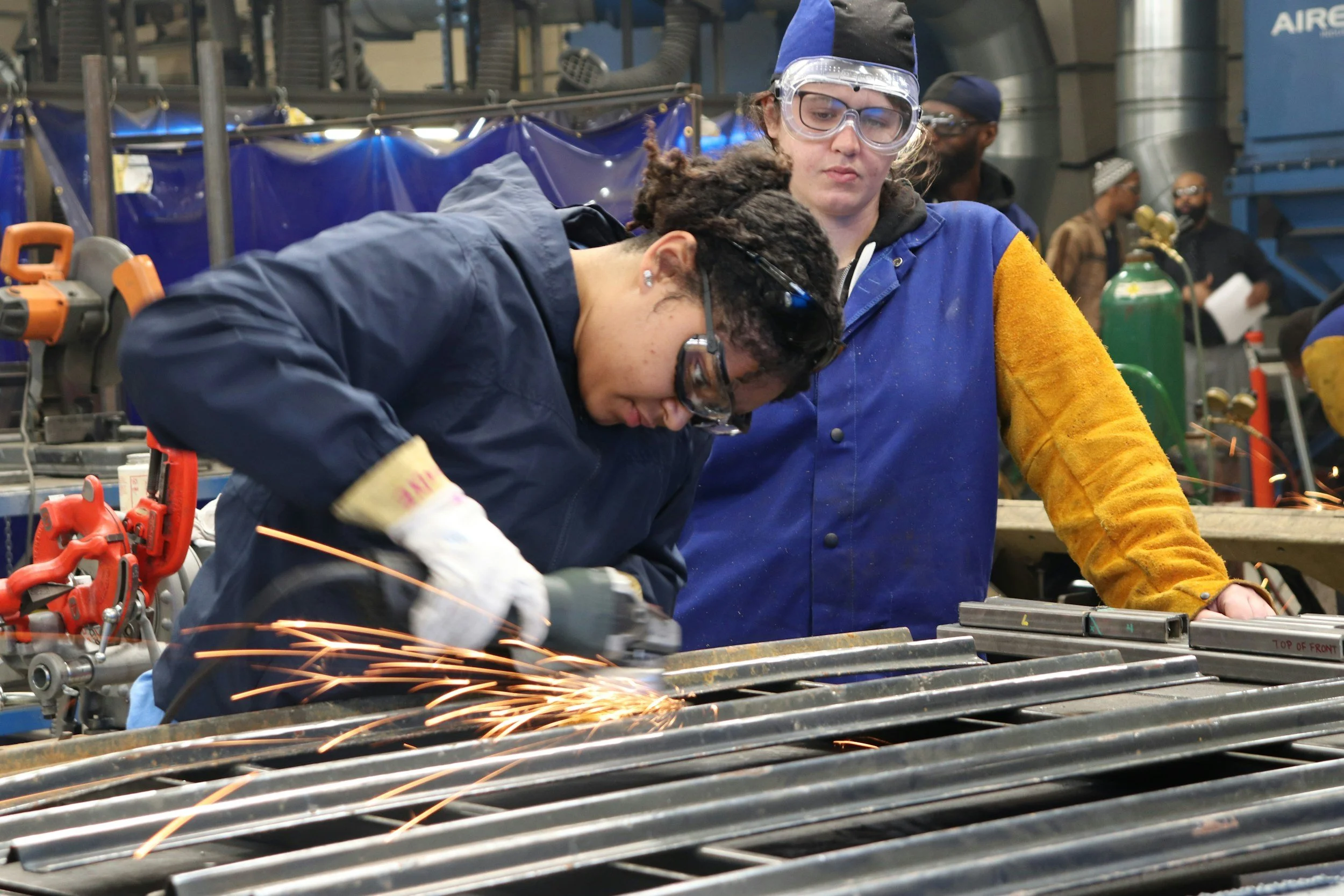 Two women working together welding metal in a factory or workshop setting, with sparks flying from the welding process.