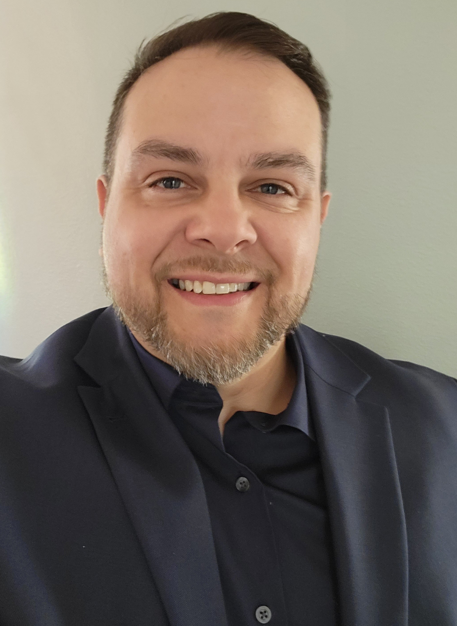 A smiling man with light skin, short brown hair, a beard, wearing a dark blazer and shirt, against a plain background.
