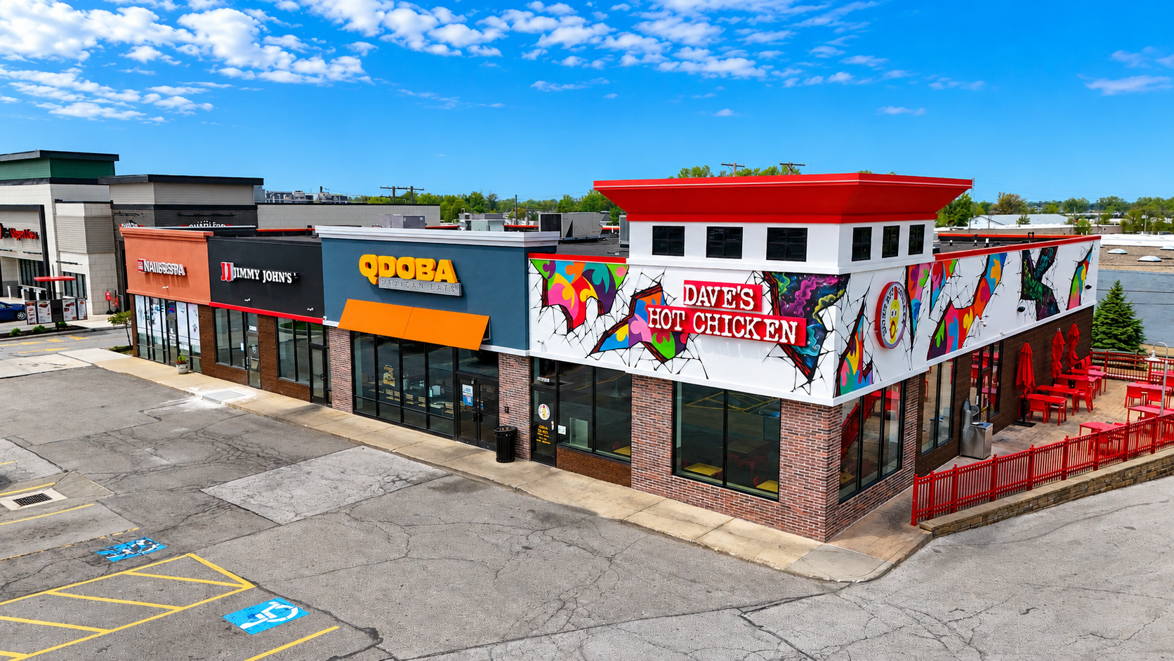 A commercial building with colorful, graffiti-style art on the upper facade and red accents, housing DAVE'S HOT CHICKEN with outdoor seating area with red umbrellas.