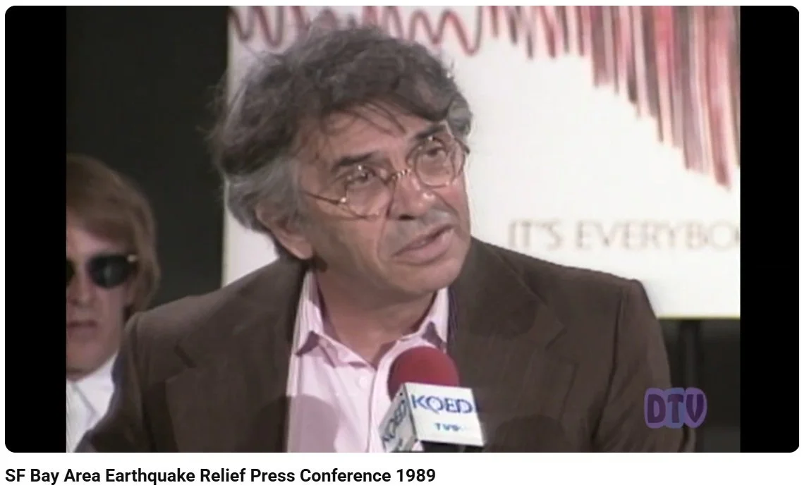 A man with gray hair and glasses speaking into a microphone at a press conference, with another person wearing sunglasses behind him, during the SF Bay Area Earthquake Relief Press Conference in 1989.
