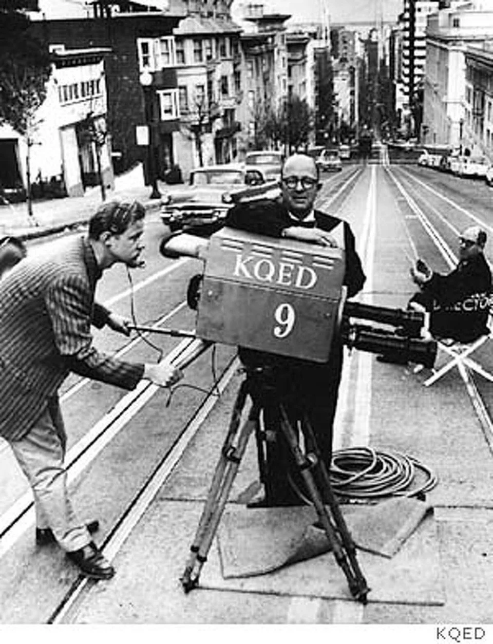 Black and white photo of a man in a tuxedo operating a vintage film camera on a city street, with buildings, parked cars, and another person in the background.