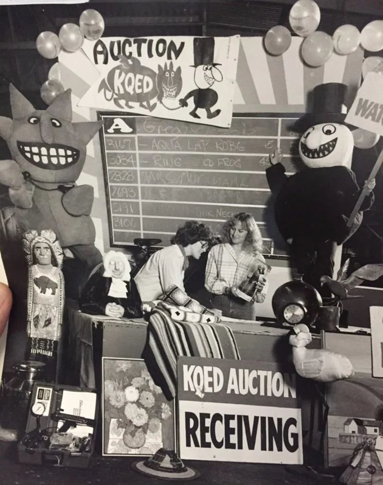 A lively auction scene with inflatable animals, including a large cat and a snowman, and a blackboard listing bids. People are engaging with the event, surrounded by balloons and decorative items.