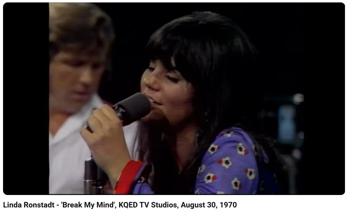 A woman with long dark hair singing into a microphone, with a man in the background, in a recording studio or TV studio setting.