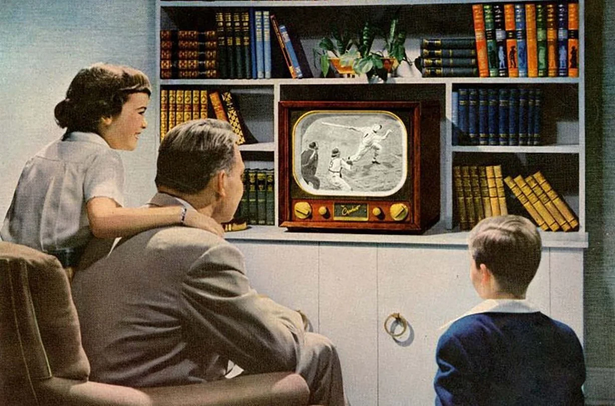 A family of four watching television in a living room, with a built-in bookshelf filled with colorful books. The TV displays a black-and-white cartoon of people running and jumping.