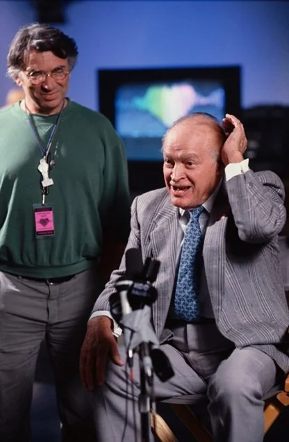 Two men having a conversation in a television studio, one seated with a grey suit and the other standing in a green sweatshirt and glasses.