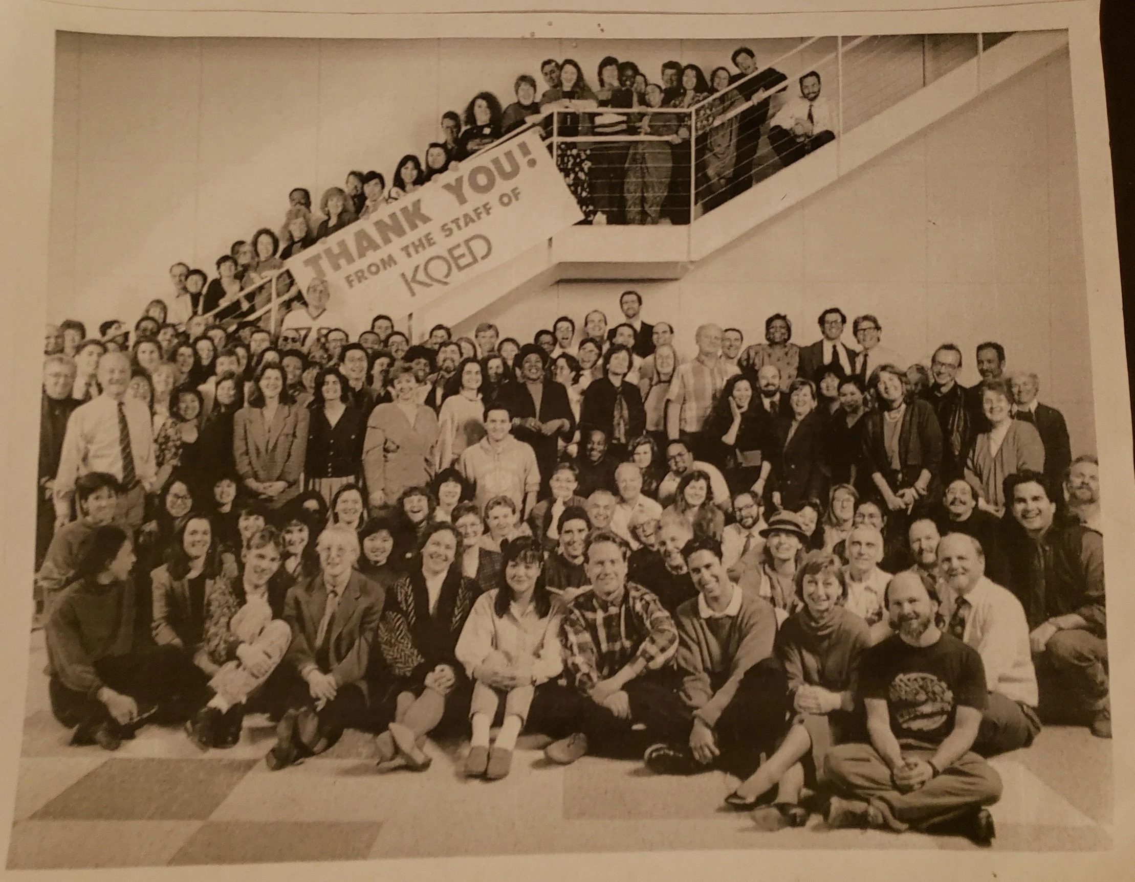 Black and white group photo of many people gathered together on stairs, with a banner reading 'Thank You! from the Staff of KOED' hanging on the railing.