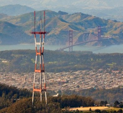 A large red and white transmission tower in the foreground with a bridge and mountains in the background.