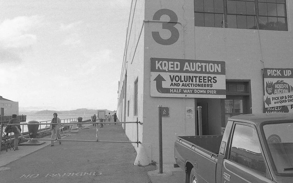 People at an outdoor pier area next to a building with signs for a KQED auction, volunteers, and pickups, with the water and hills in the background.