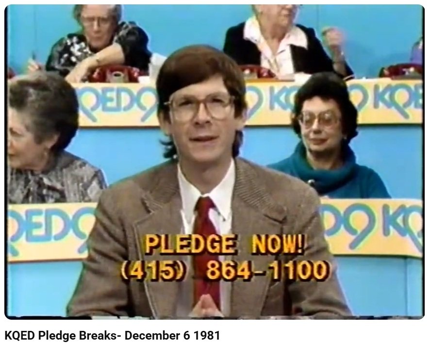 A man in a gray suit with glasses in a TV studio with other elderly people with pastel blue background and colorful signs reading 'KQED'. The man is speaking, and text in yellow and black at the bottom reads 'PLEDGE NOW! (415) 864-1100'.