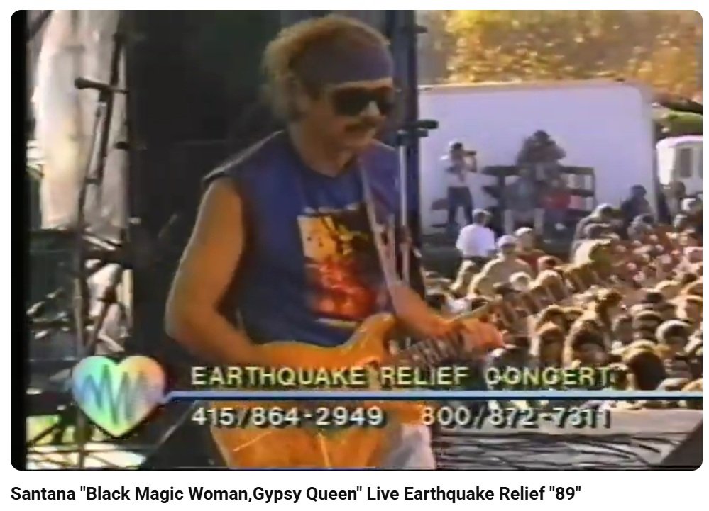 A woman wearing sunglasses and a colorful shirt playing an electric guitar at an outdoor earthquake relief concert with a crowd in the background.