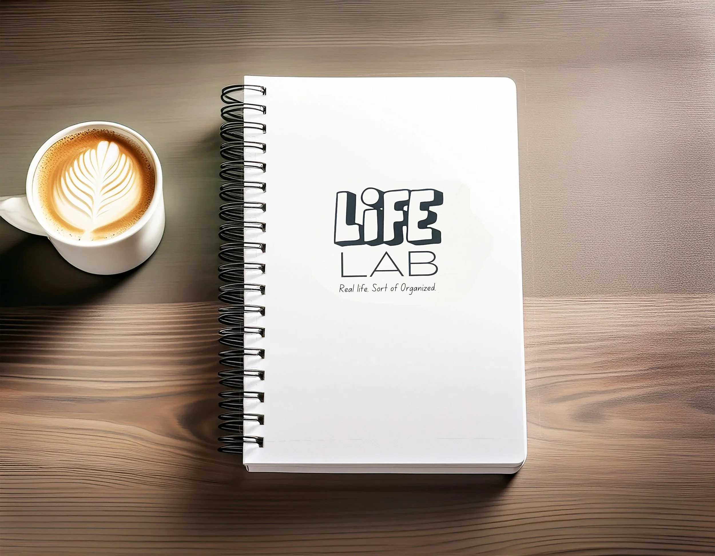 A notebook titled 'LIFE LAB' with a subtitle 'Real life. Sort of Organized.' on a wooden desk, next to a white coffee mug with latte art in the shape of a leaf.