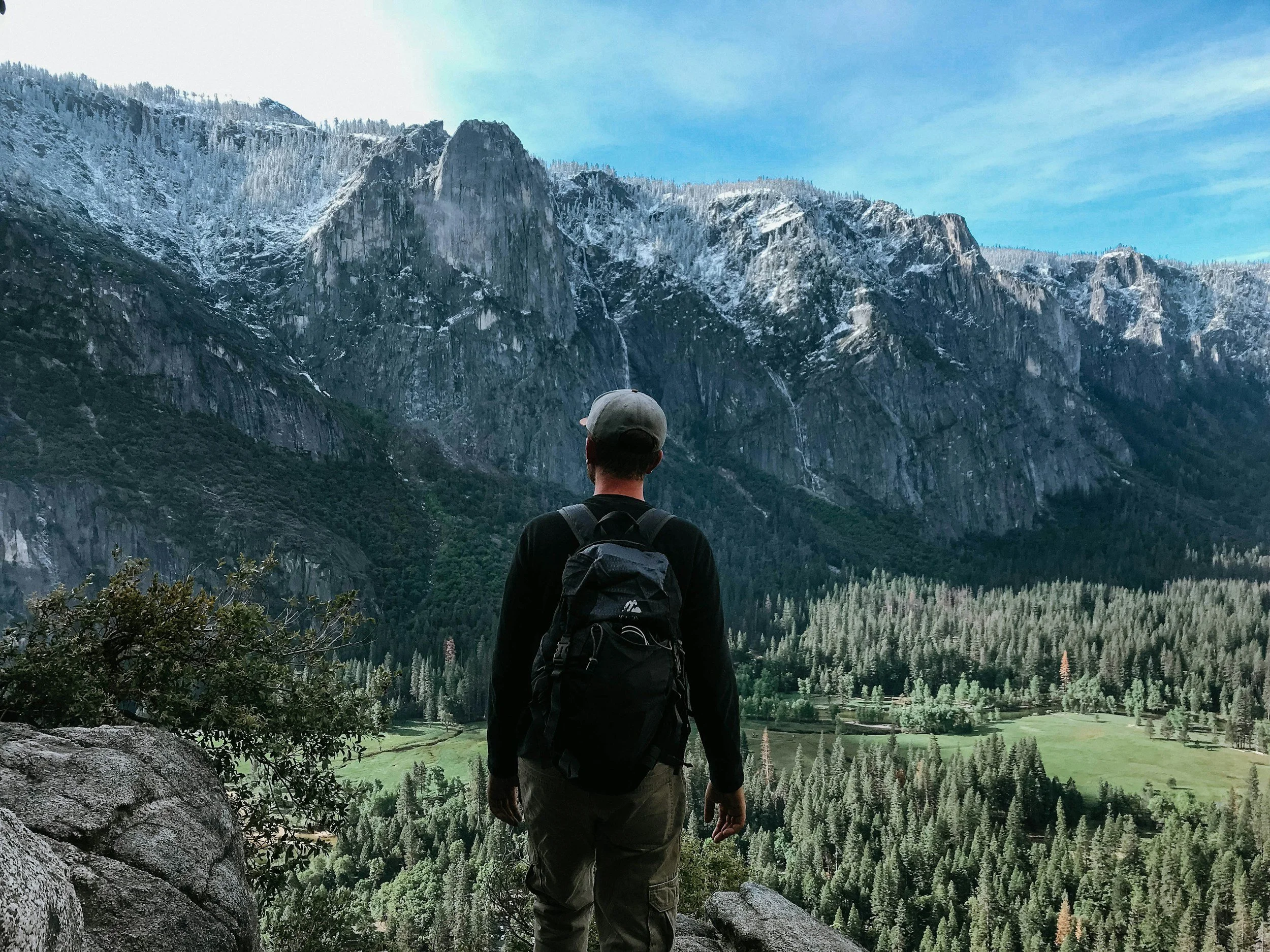 A person with a backpack standing on a rocky ledge, looking at a valley surrounded by tall, snow-capped mountains and dense forest.
