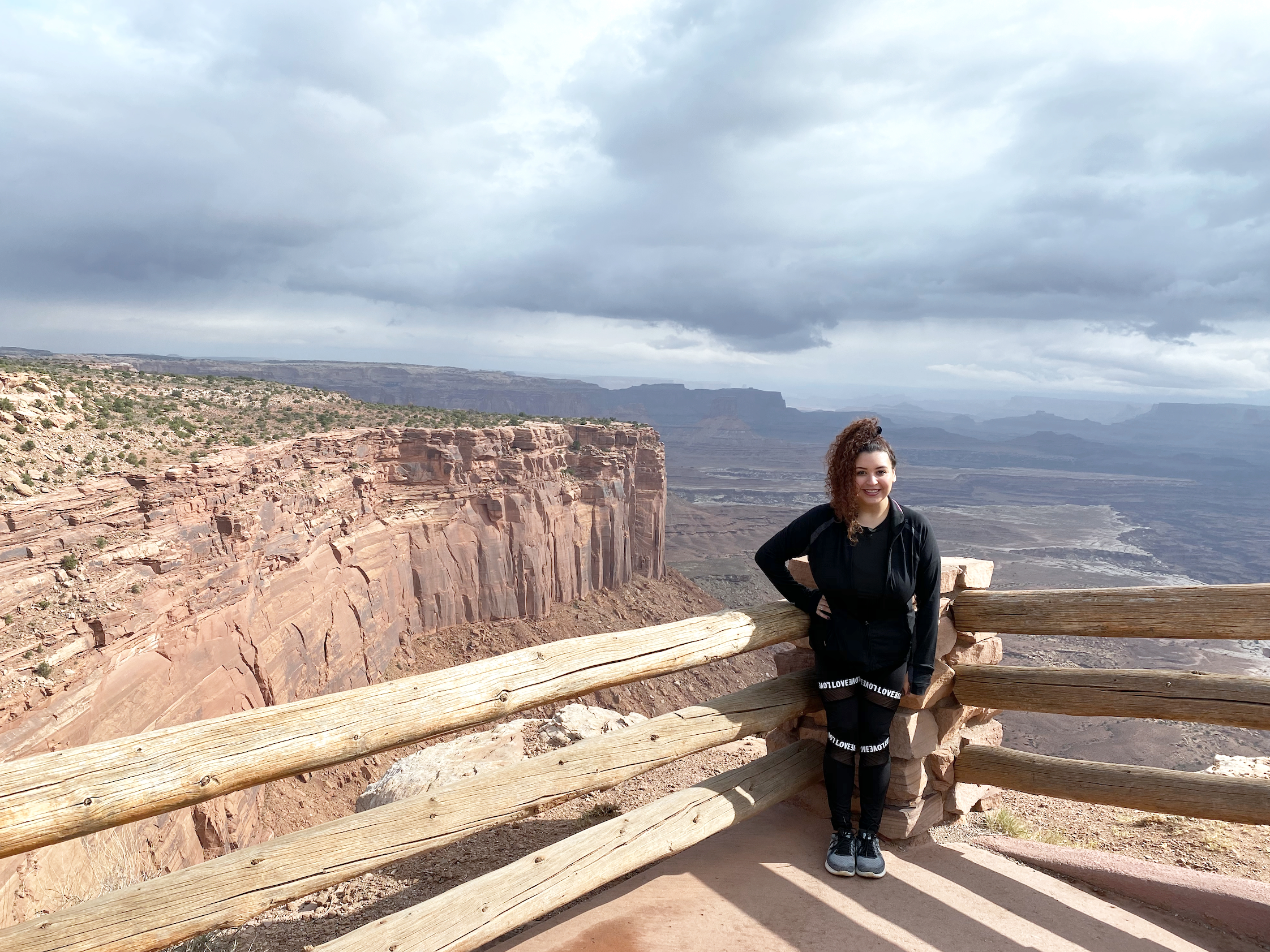 A woman standing by a wooden railing with a scenic canyon landscape and cloudy sky in the background.