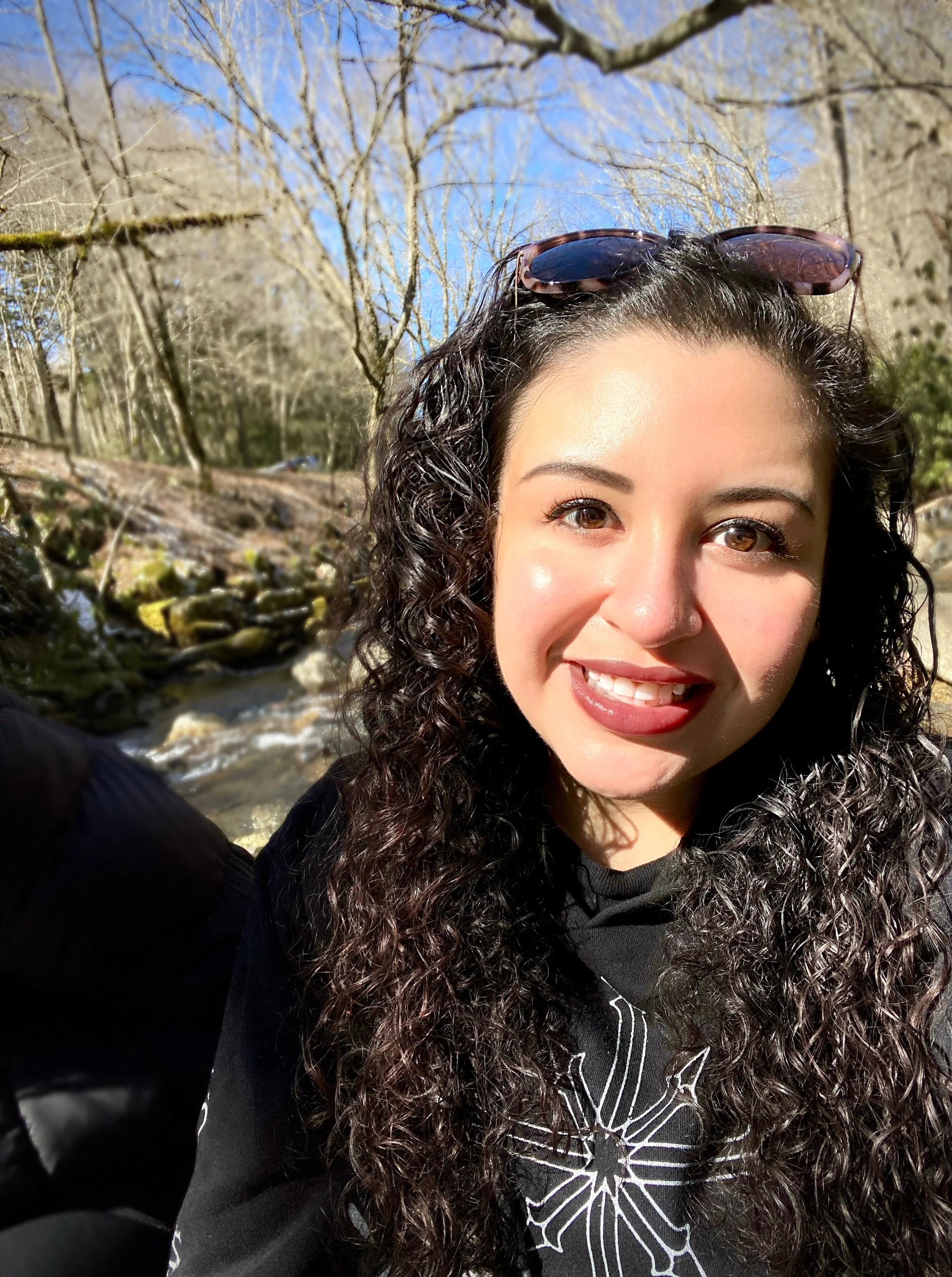A young woman with curly hair and sunglasses on her head smiling outdoors near a creek with trees in the background.