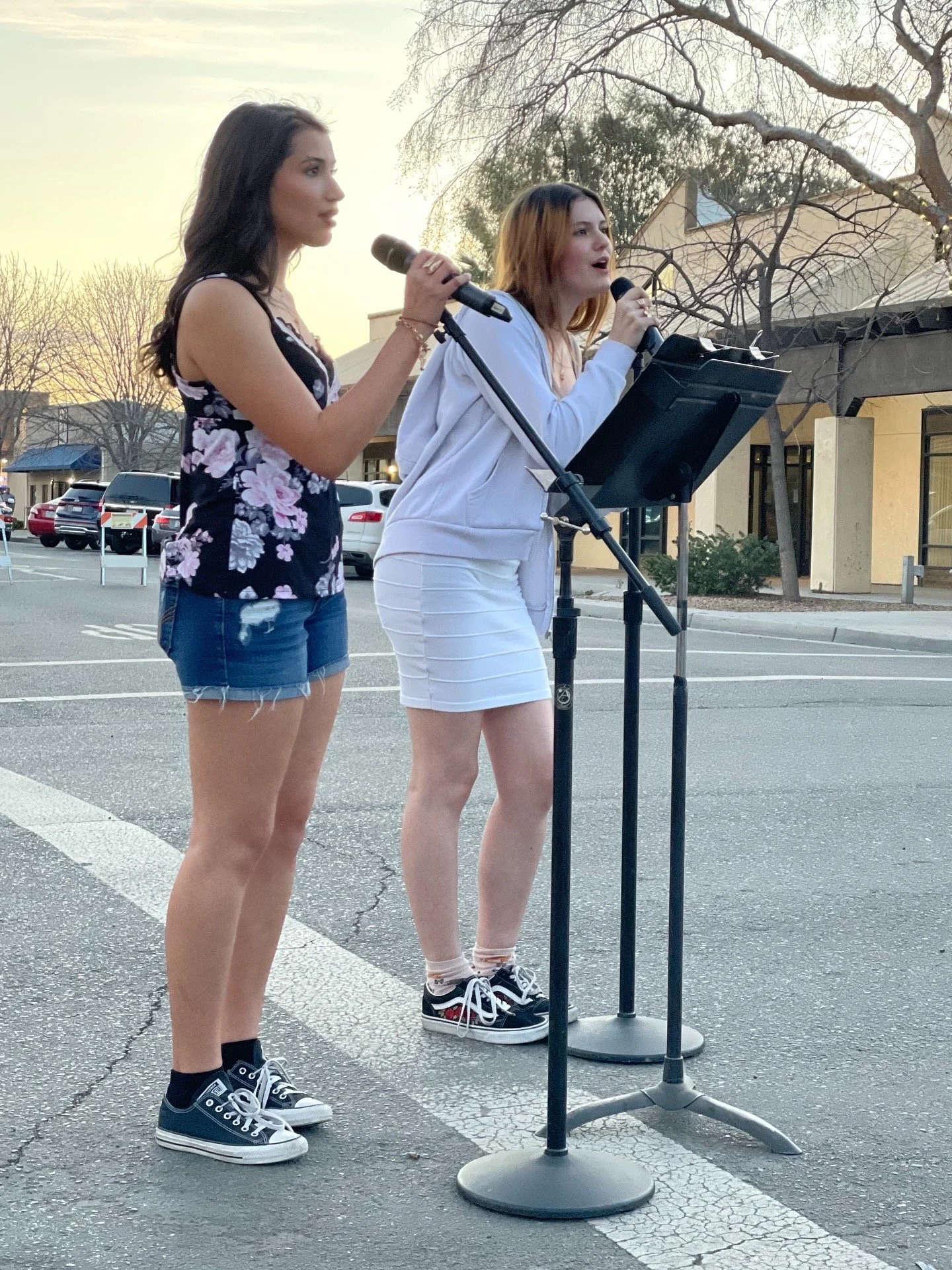 Two young women singing into microphones downtown in a small town during sunset, standing next to a music stand with a music sheet, in a parking lot with bare trees and buildings in the background. Mardi Gras event in Coalinga, California.