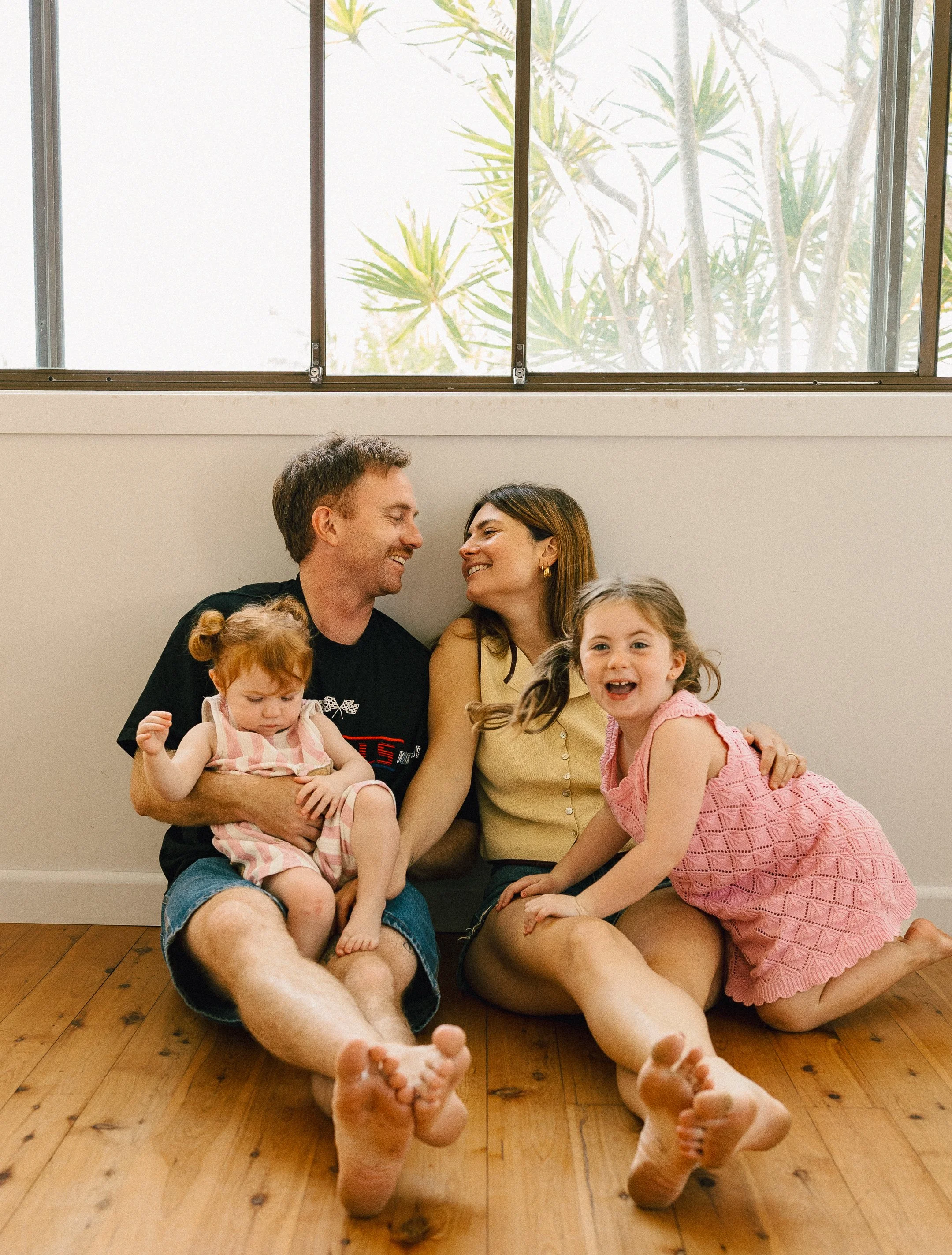 A happy family sitting on a wooden floor inside a home, face to face, smiling at each other, with large windows and leafy plants visible in the background.