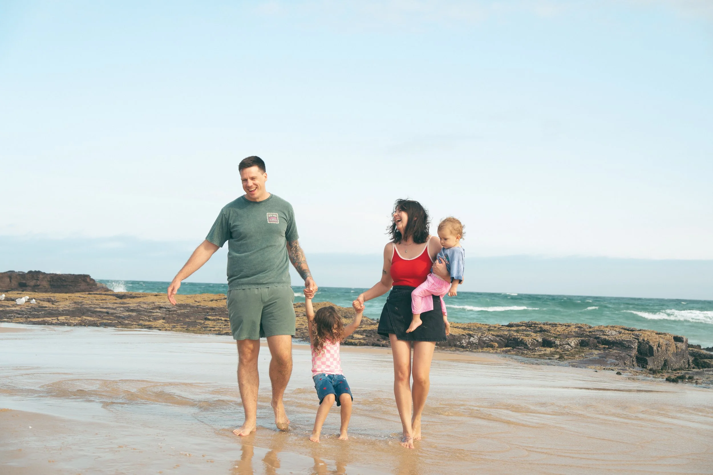 A family of five holding hands and walking at the beach, with the ocean, rocks, and sky in the background. The parents are smiling and children are playing.