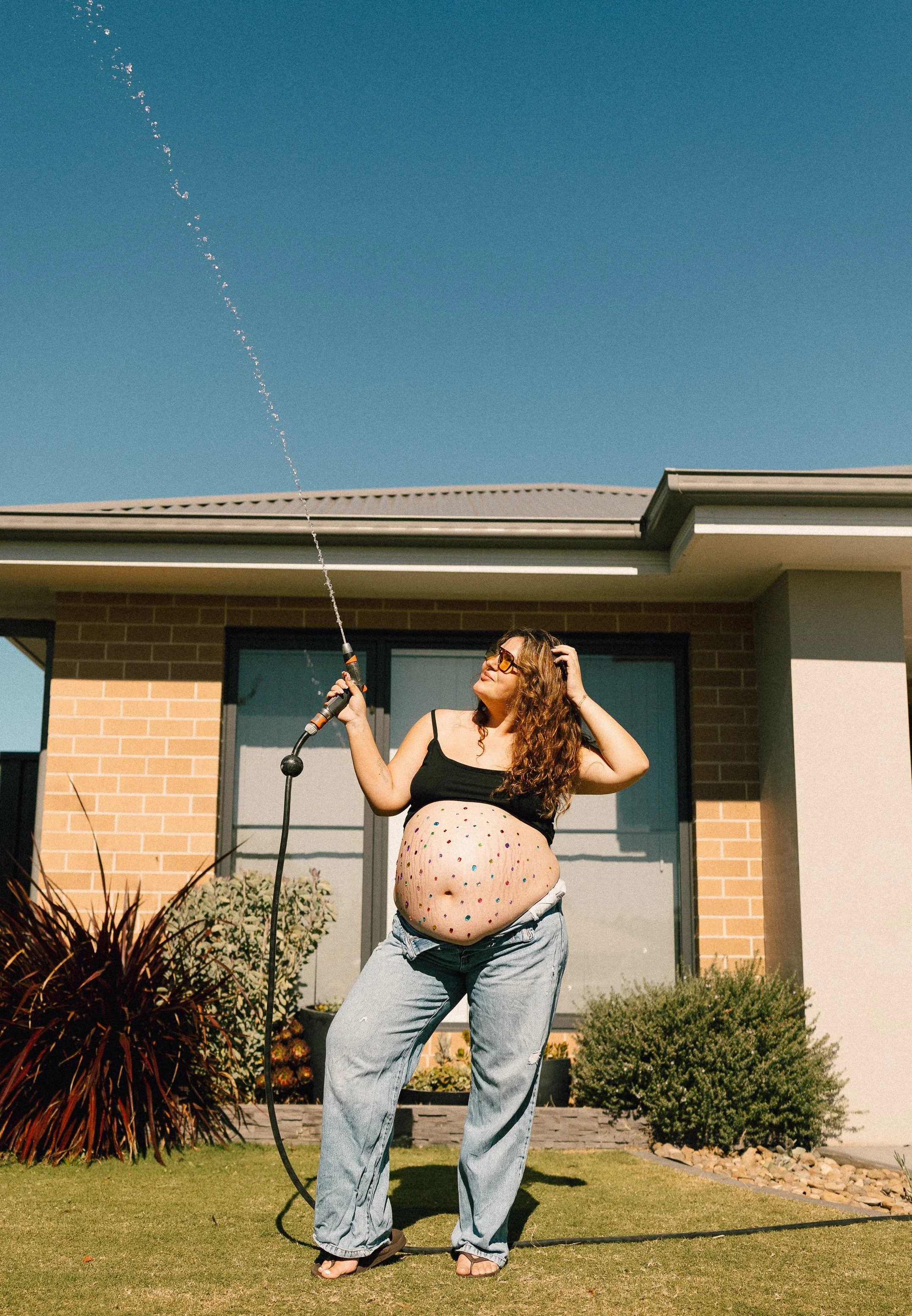 A woman with curly hair wearing glasses, a black tank top, and jeans is standing on a lawn with her pregnant belly exposed and painted with colorful dots. She is holding a garden hose spraying water into the air.