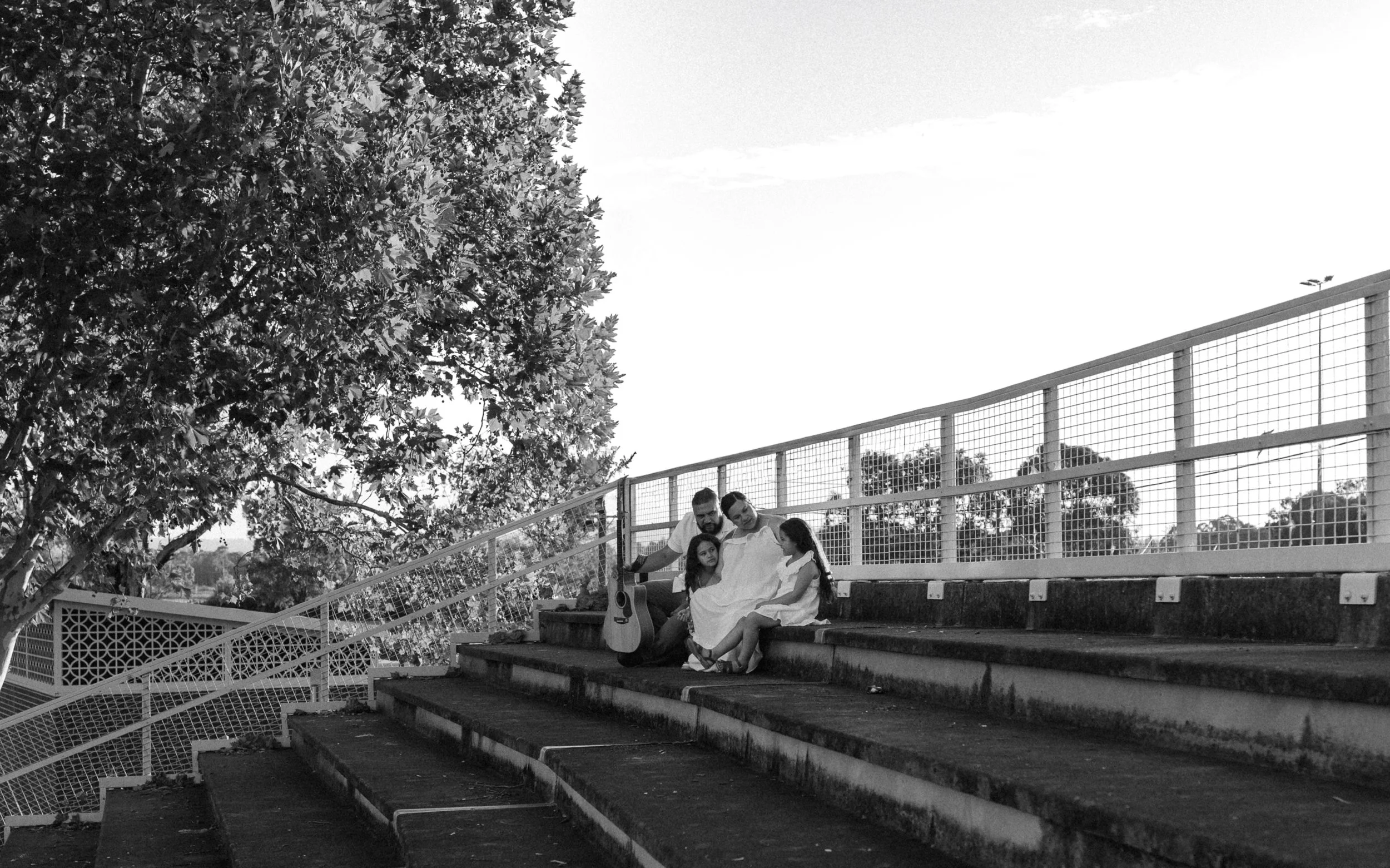 A family of four sitting on outdoor stadium steps, with a guitar resting on their laps. They appear to be enjoying each other's company, with a large tree and a sports field fencing in the background.