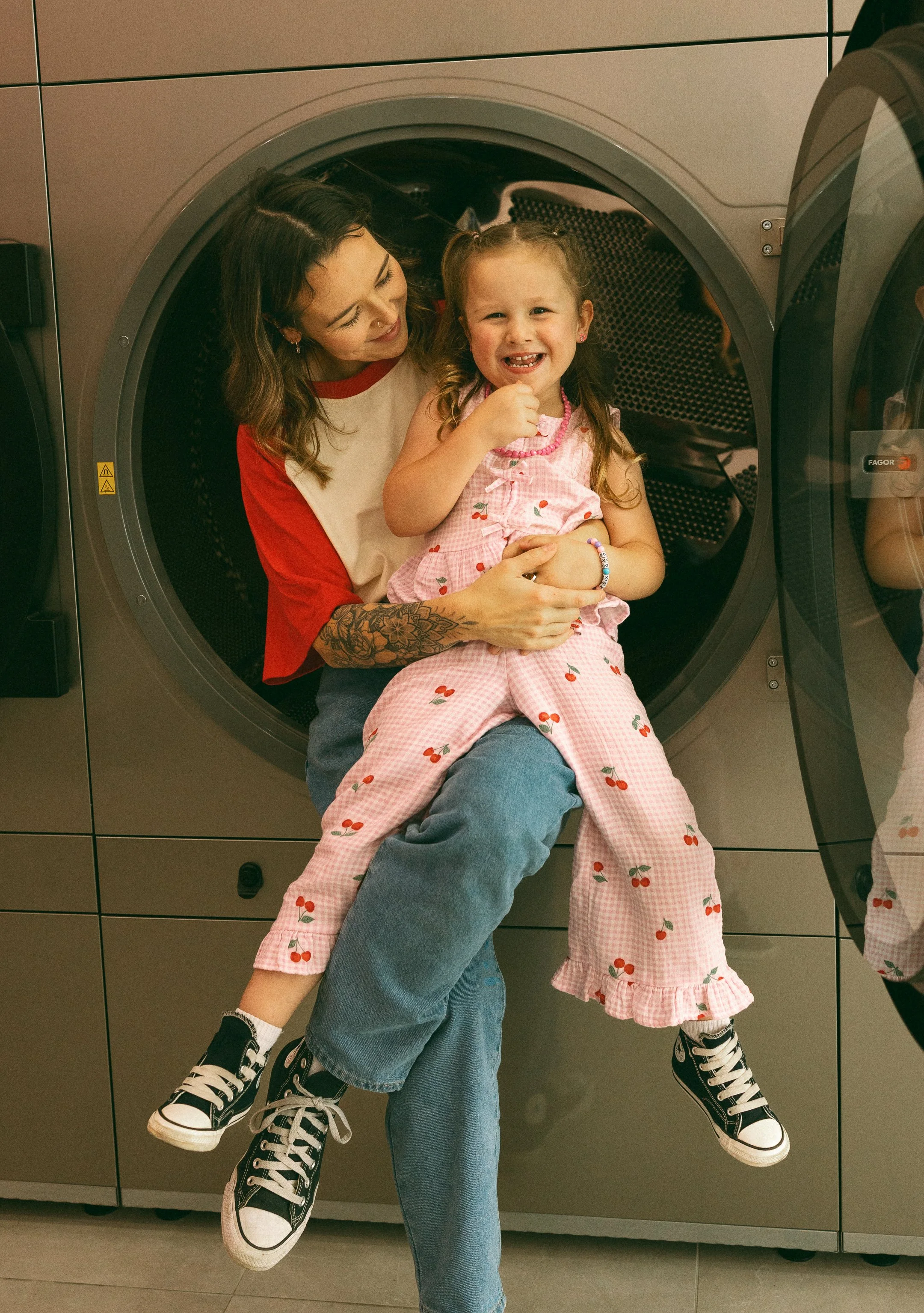 A woman with tattoos on her arm holding a smiling young girl with pigtails and a pink cherry-patterned dress sitting on her lap inside a laundry room.