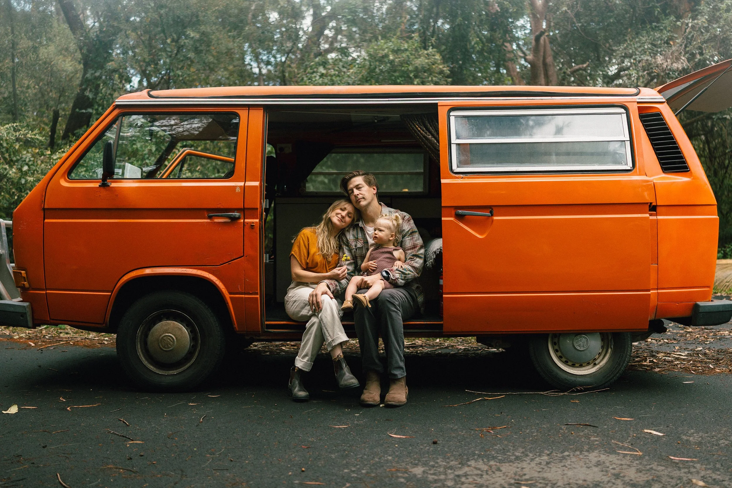 A family of three sitting inside an orange vintage van with the sliding door open, parked in a forested area. The woman is leaning on the man's shoulder, holding a small bouquet, while the young child sits on the man's lap. The man has his eyes closed, appearing relaxed, and the child looks to the side. The van has a large window with curtains, and the background shows trees with green foliage.