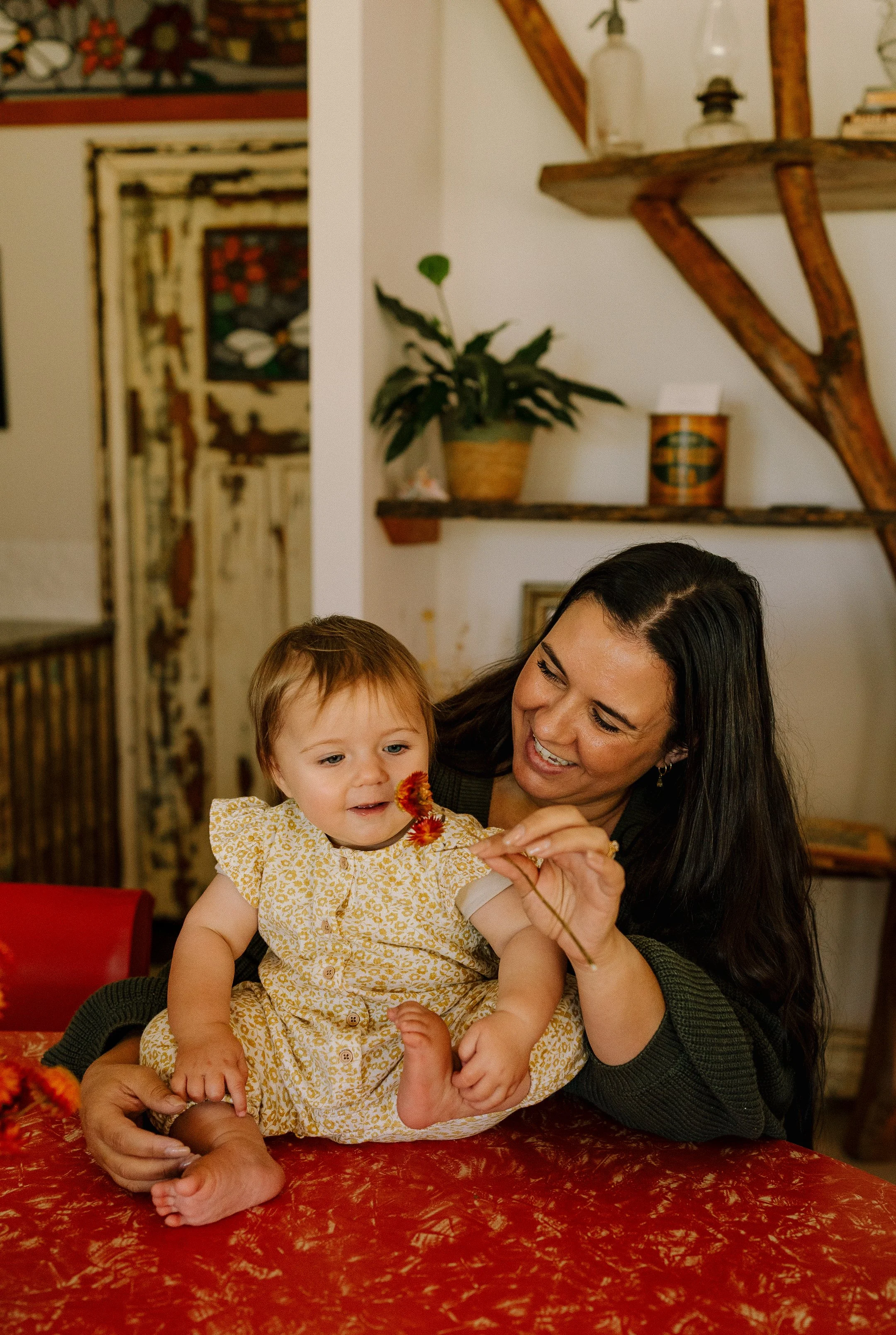 A woman with black hair and a woman with light brown hair hold a child with red hair, and the woman with black hair helps the child hold a flower, sitting at a table covered with a red patterned tablecloth in a cozy room.