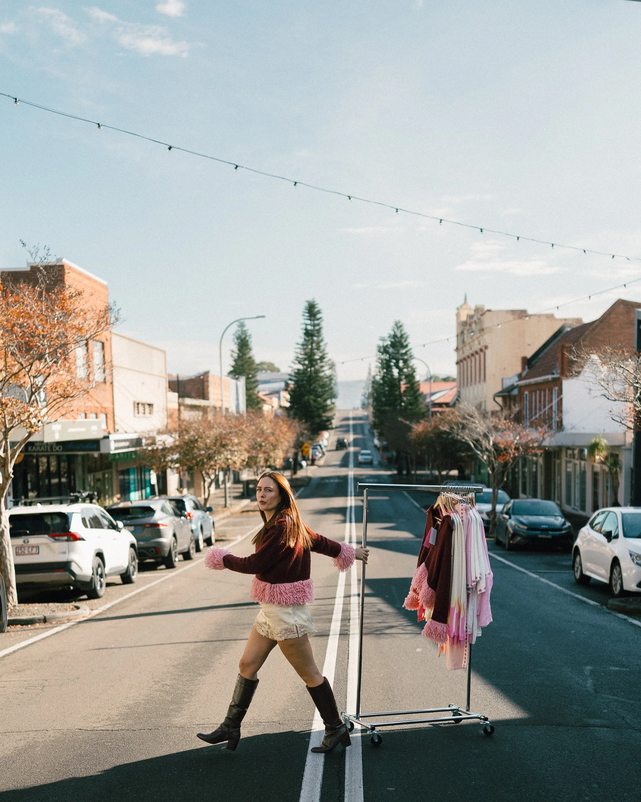 A young woman with long brown hair wearing a fluffy pink and burgundy sweater and beige shorts walks across a city street while pushing a clothing rack with pink and beige dresses. There are parked cars and buildings on both sides of the street, with a few trees and a partly cloudy sky.