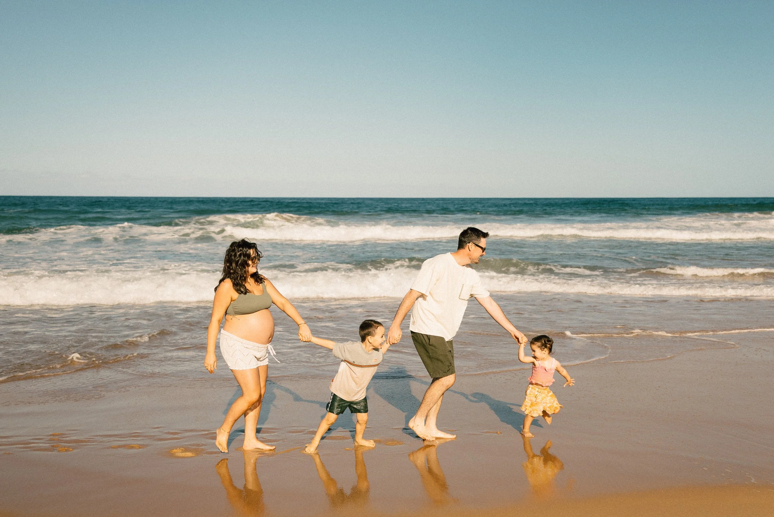 A family walking hand in hand on the beach. The woman has a pregnant belly and curly dark hair. The man wearing glasses and a white shirt. Two young children, one boy and one girl, holding hands with the adults. The ocean waves in the background and clear sky.