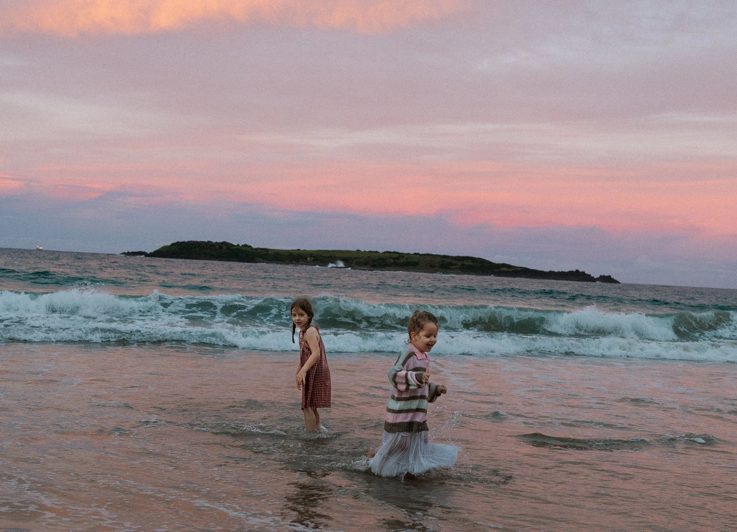 Three children playing in the ocean at sunset with pink and purple sky and an island in the background.