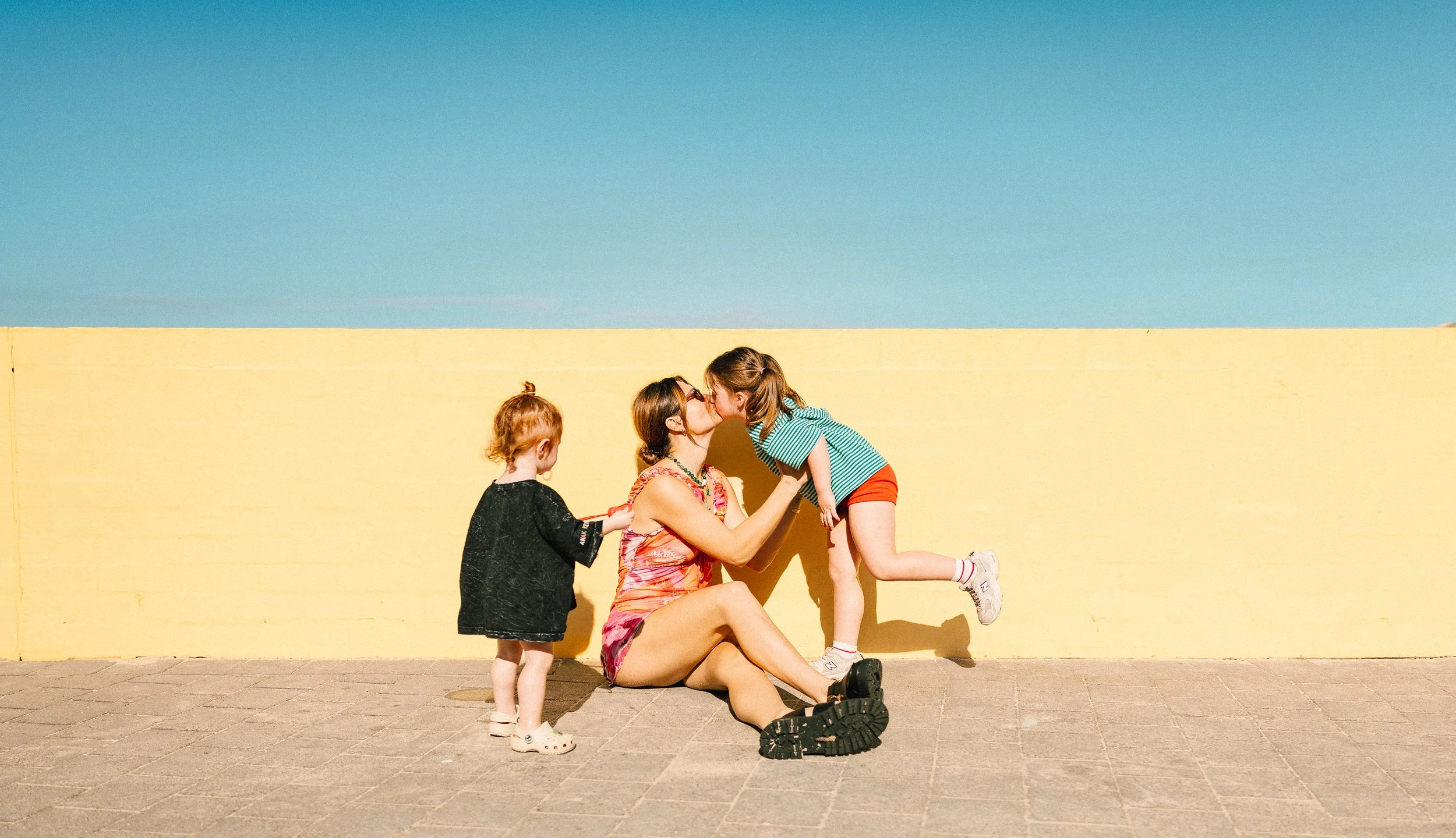 A woman sitting on the ground leaning against a yellow wall, kissing a young girl who is standing with one foot raised and leaning towards her. Two other children, one with red hair and a black jacket and another with glasses and a striped shirt, are nearby in front of the woman.